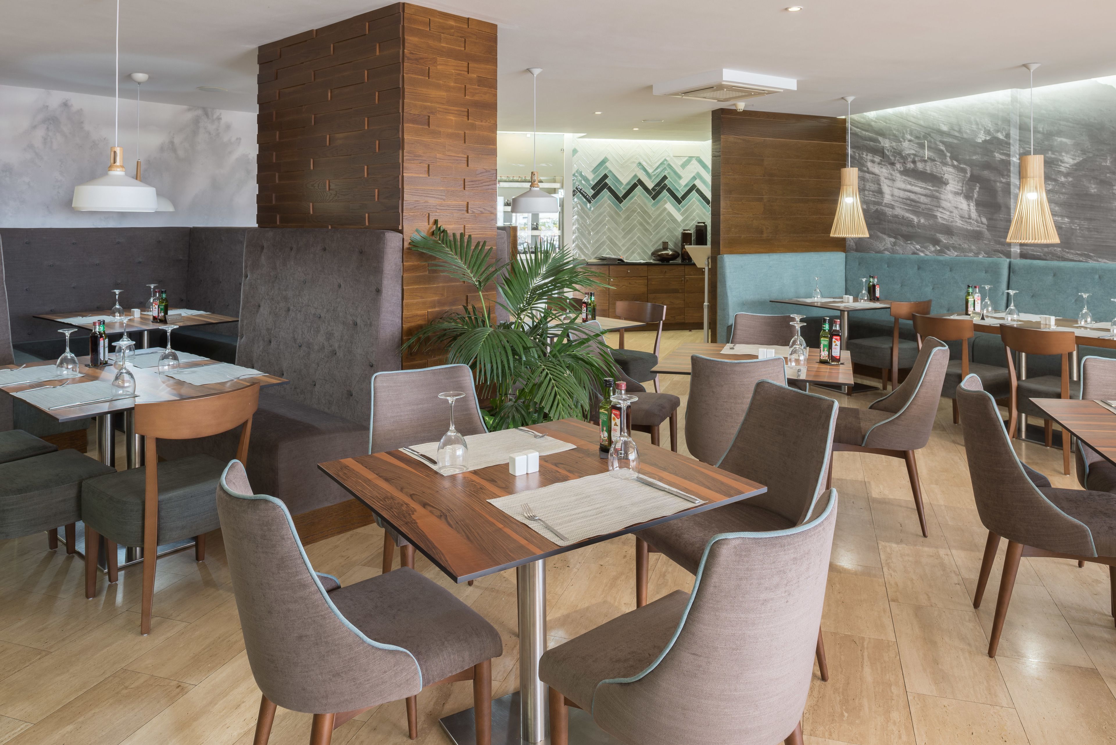 A modern restaurant with wooden tables and grey upholstered chairs, a large potted plant, and a chevron-patterned wall.