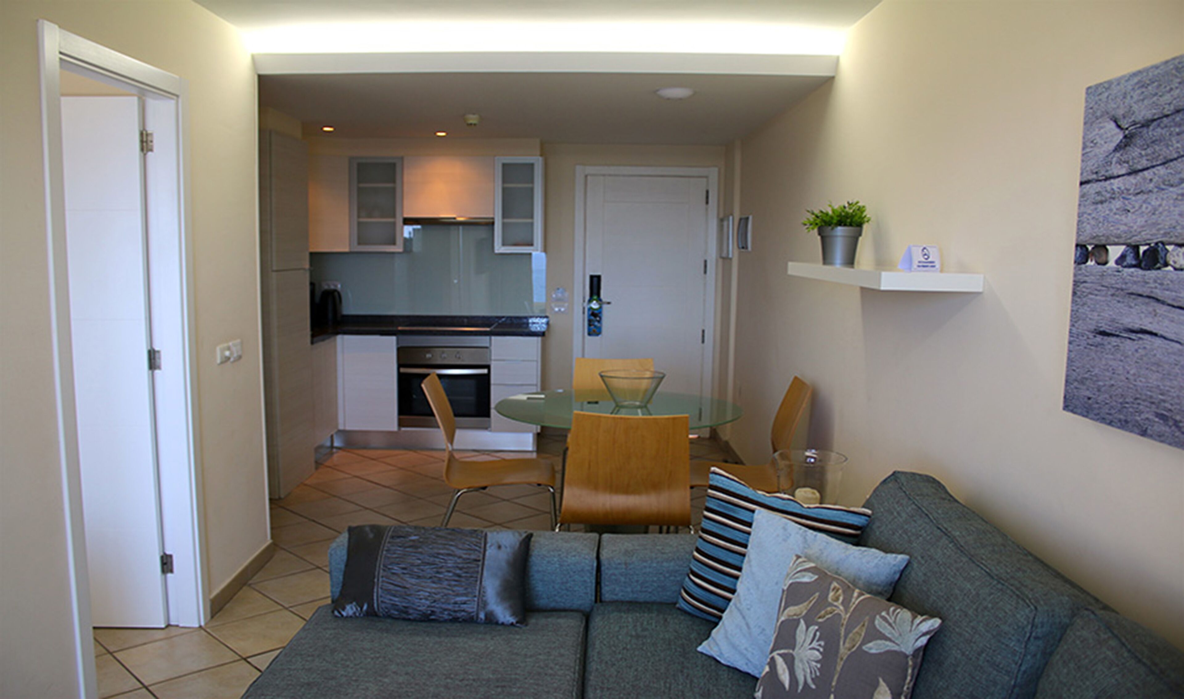 Un espacio habitable compacto con kitchenette, una zona de comedor con mesa y sillas de vidrio, y un sofá con cojines.