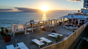 Eine Dachterrasse mit Liegestühlen, Sonnenschirmen und Topfpflanzen mit Blick auf das Meer bei Sonnenuntergang.