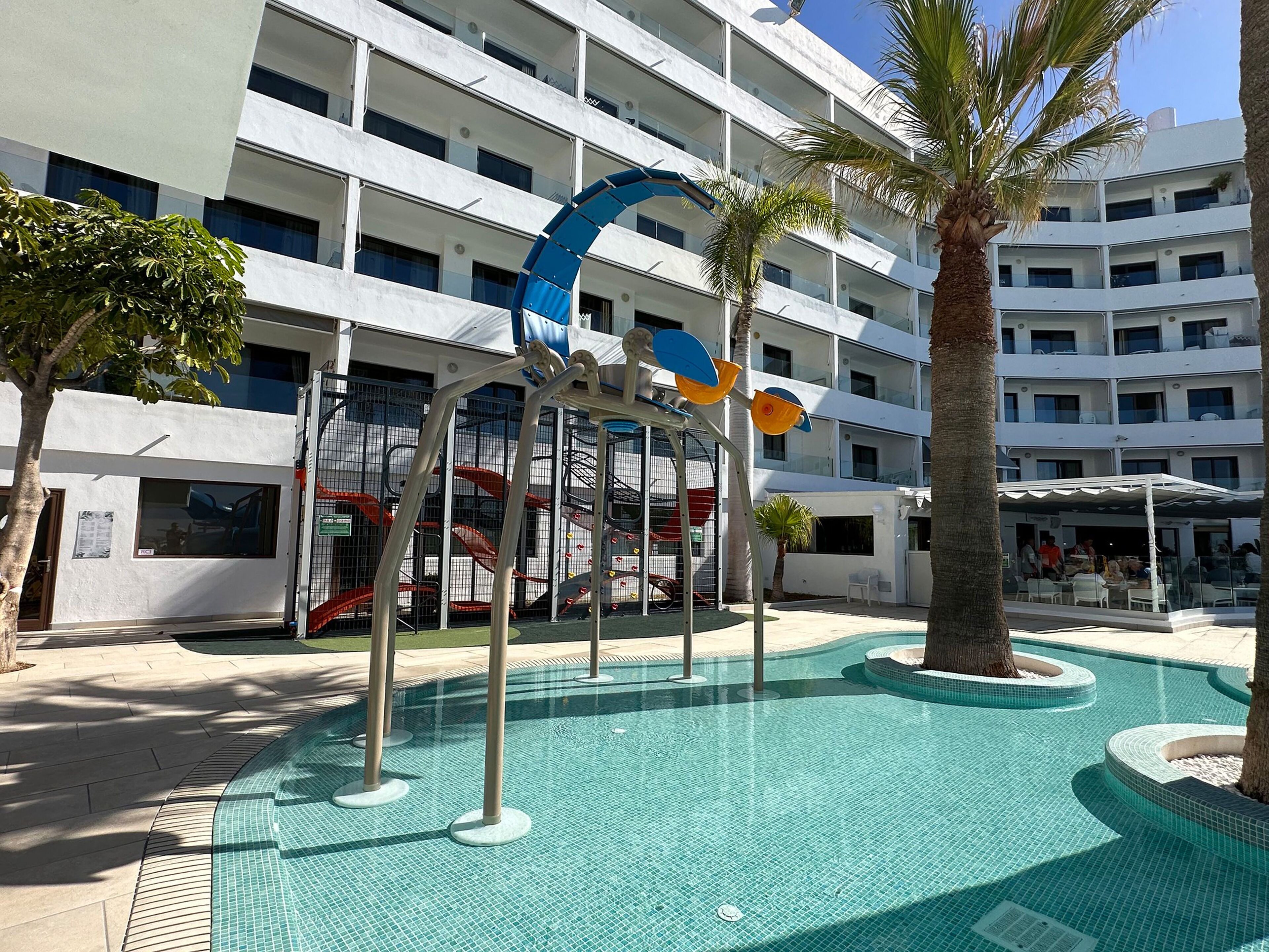 A swimming pool area with a modern building in the background, a palm tree, and a children's play structure.