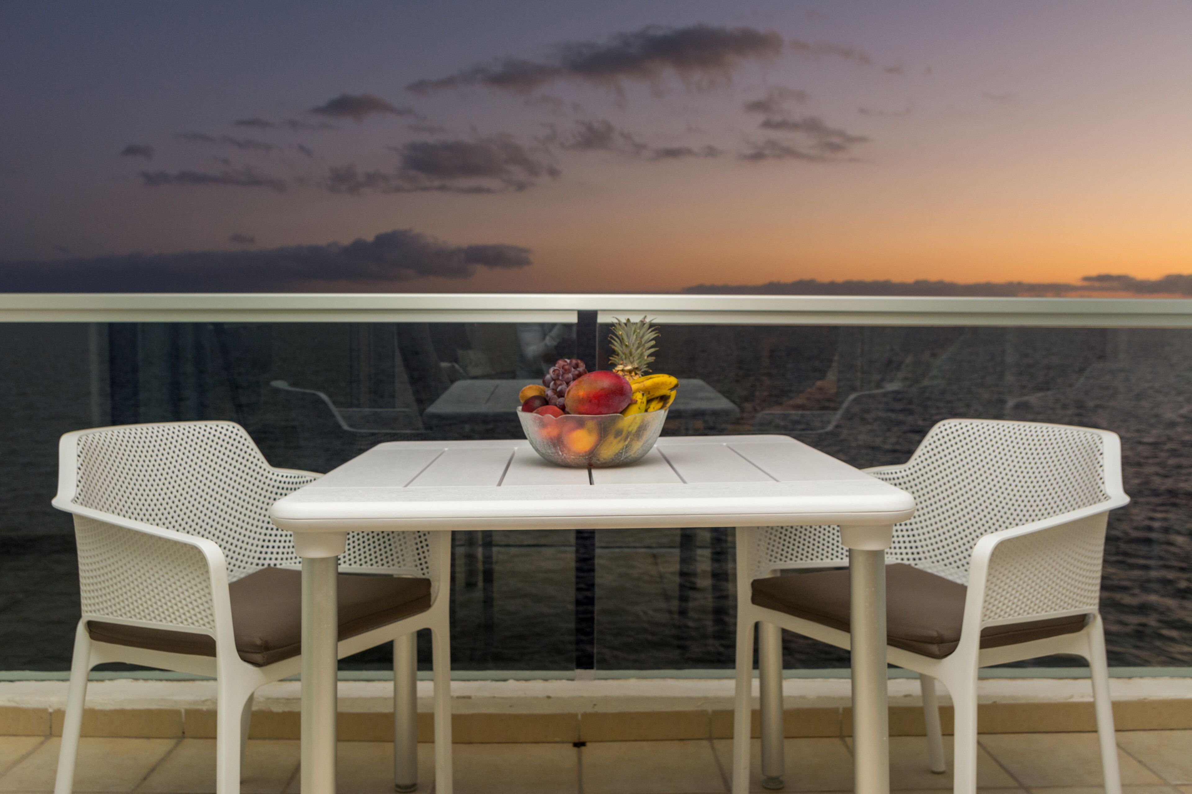 A balcony with a table and two chairs, a bowl of fruit, and a view of the ocean at sunset.