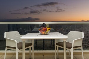 Un balcón con una mesa, dos sillas, un frutero y vistas al océano al atardecer.