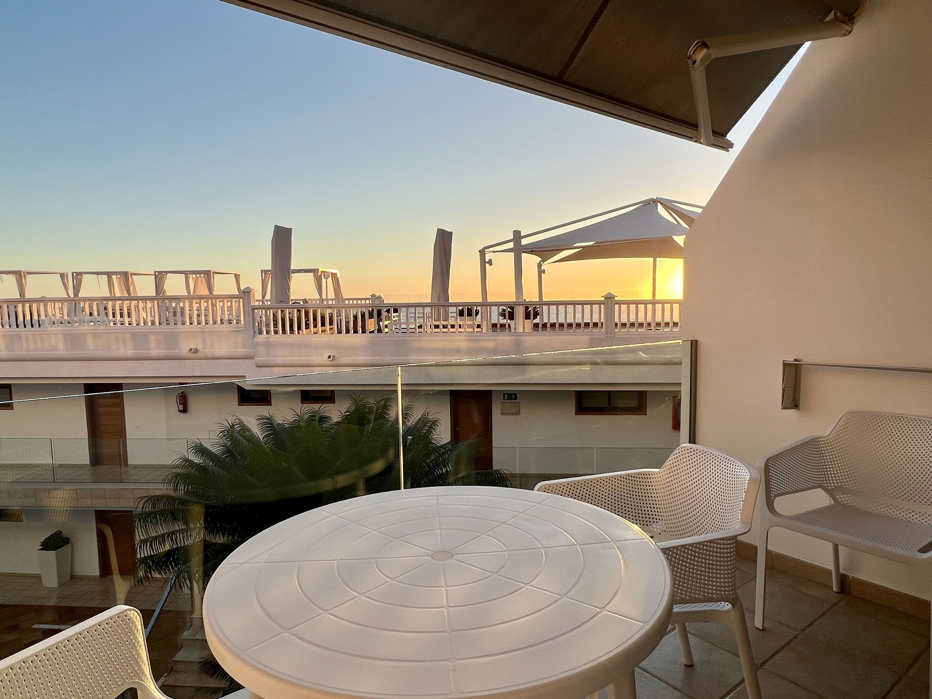 A terrace with a round table and wicker chairs, overlooking a building with a balcony and a white fence.