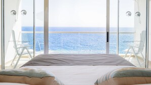 Ein Schlafzimmer mit einem großen Bett, zwei weißen Stühlen und Blick auf den Ozean durch große Glasfenster.