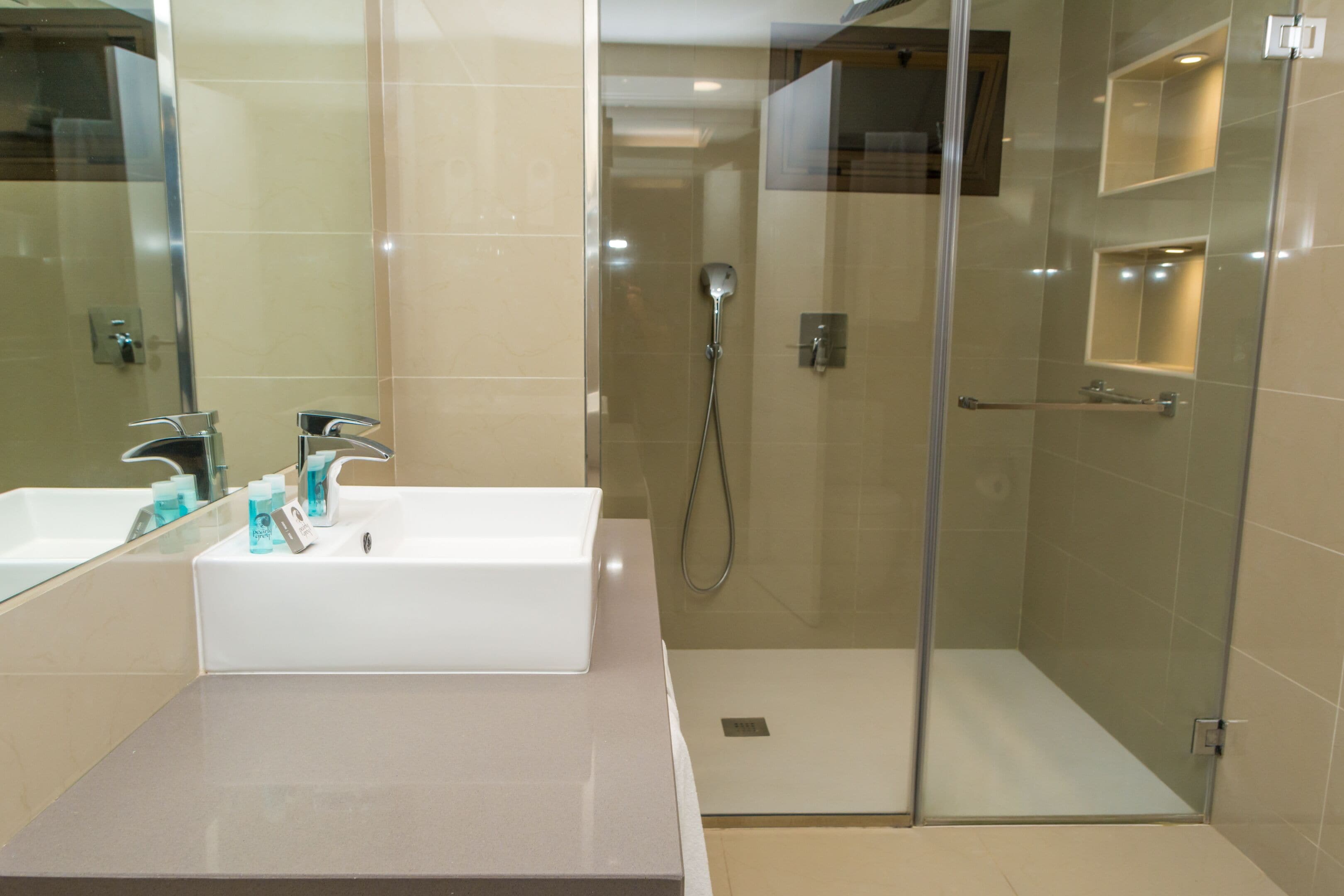 Ein modernes Badezimmer mit einer gläsernen Duschkabine, einem weißen Waschbecken mit Spiegel und einer beige gefliesten Wand.