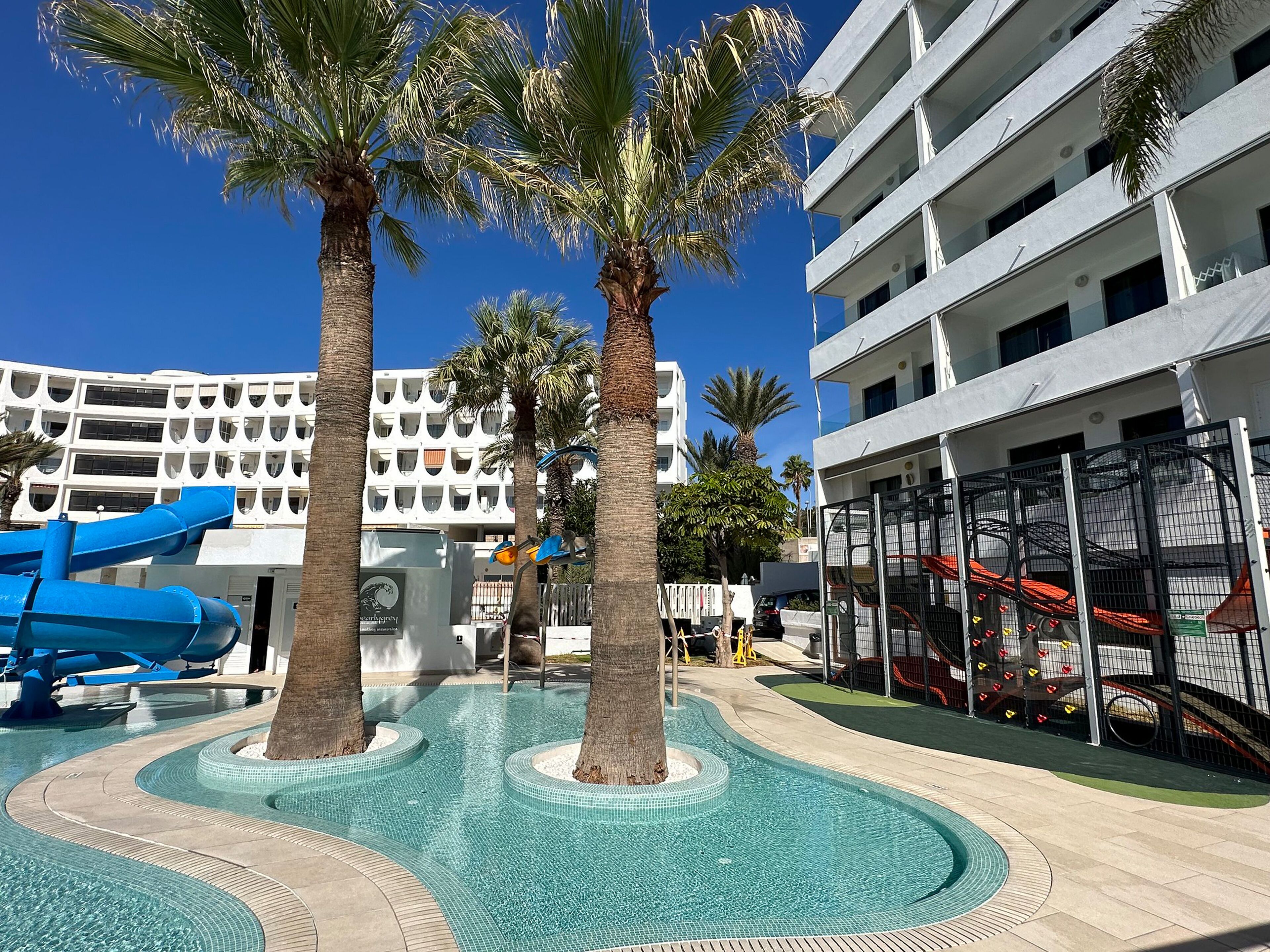 A pool area with palm trees, a blue slide, and a white building with balconies.