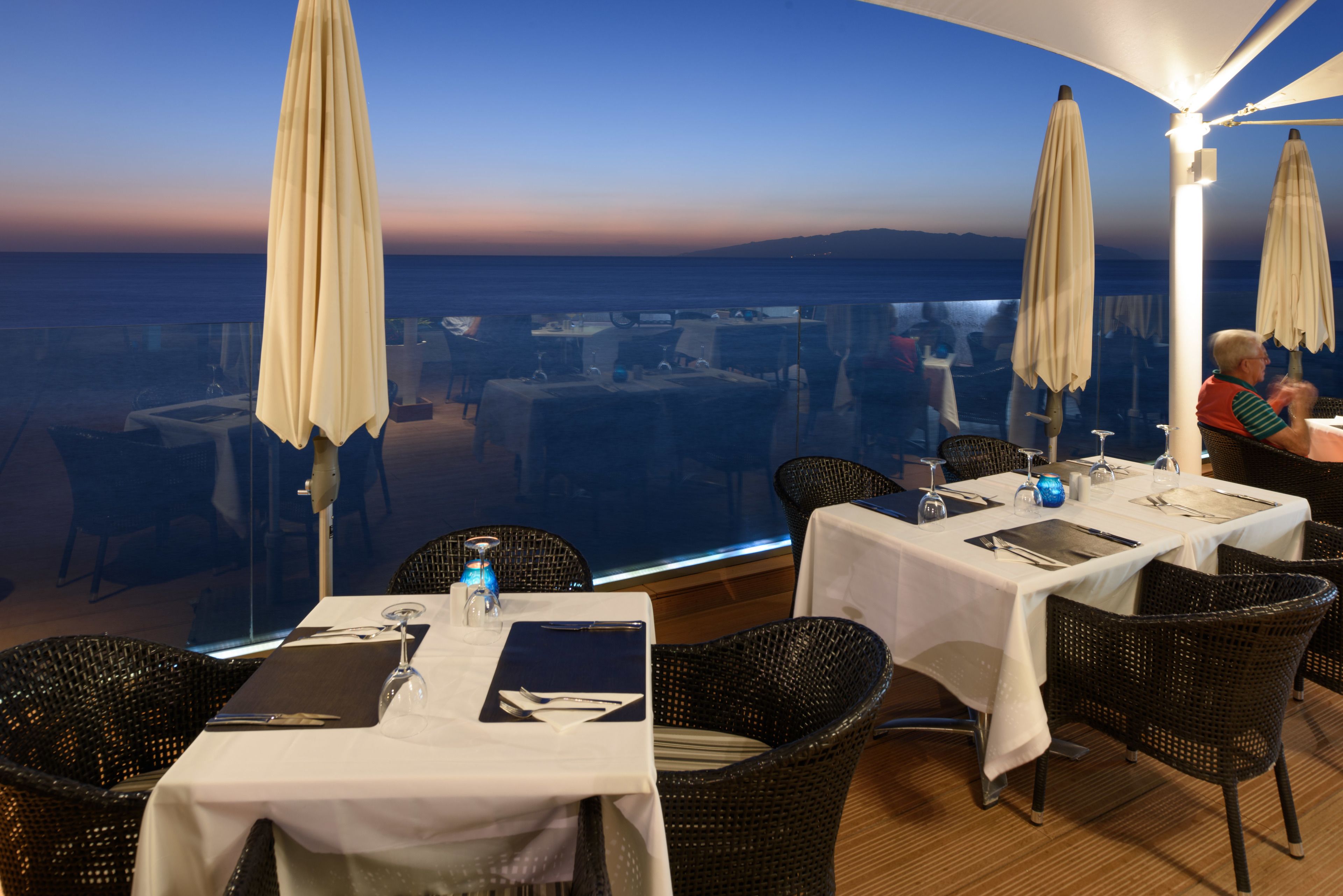 A restaurant with tables set for dining, outdoor umbrellas, and a view of the sea.