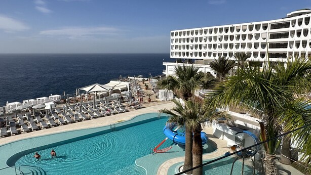 Ein Poolbereich mit Blick auf das Meer, Liegestühle und ein großes Gebäude im Hintergrund.
