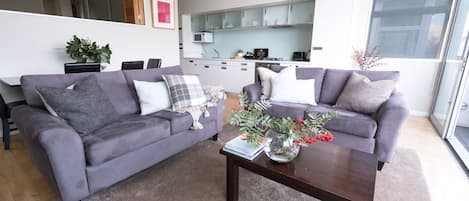 Una sala de estar moderna con dos sofás grises, una mesa de centro con un florero y una cocina al fondo.