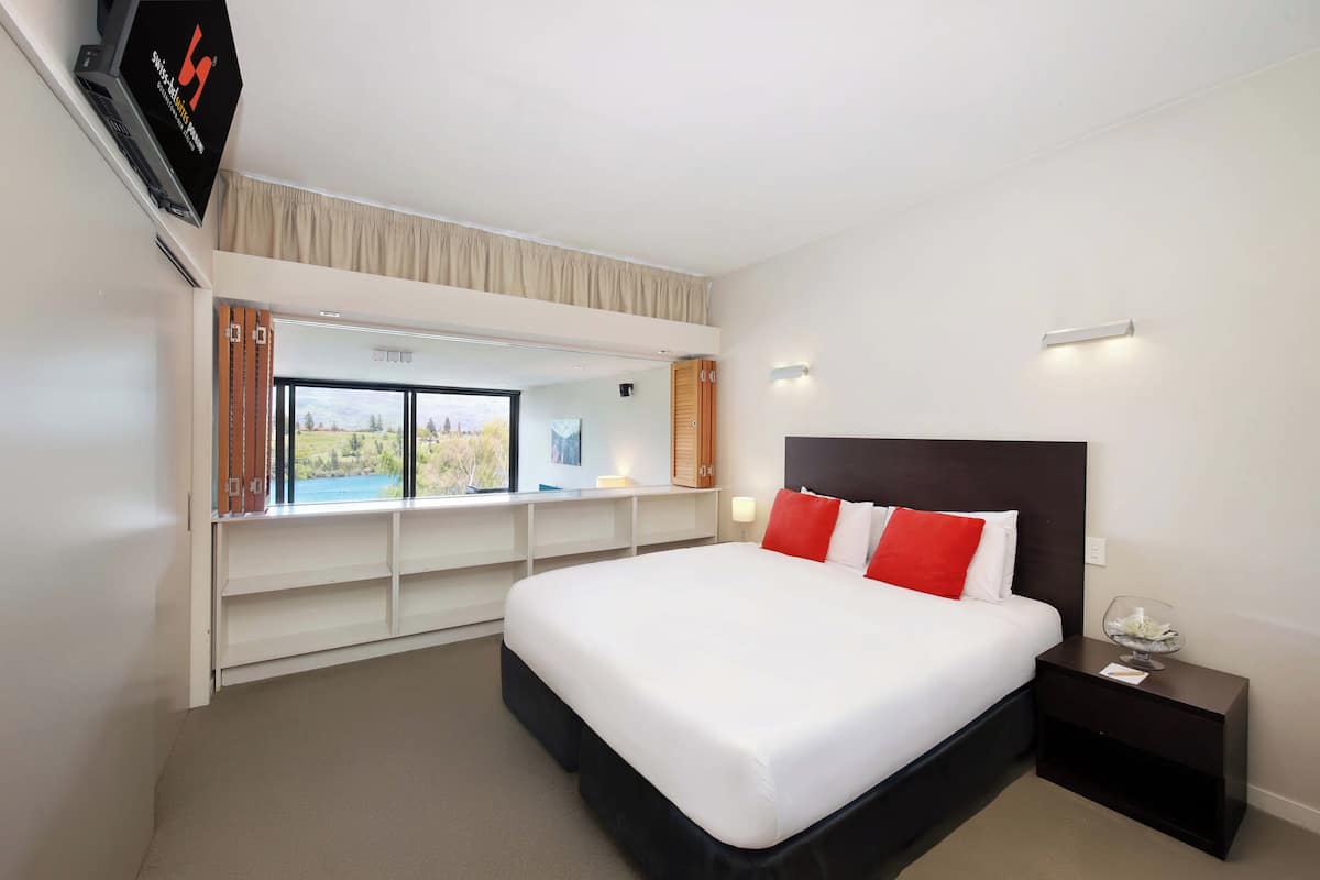 Una habitación de hotel con una cama grande, un televisor montado en la pared, una ventana con vistas y una mesita de noche con una lámpara.