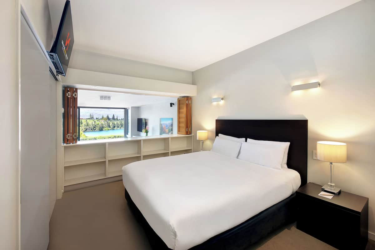 Una habitación de hotel con una cama grande, lámparas de noche, un televisor montado en la pared y vistas a un lago a través de una ventana.