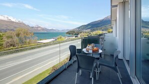 Un balcón con una mesa dispuesta para el desayuno, con vistas a una carretera y un lago con montañas al fondo.