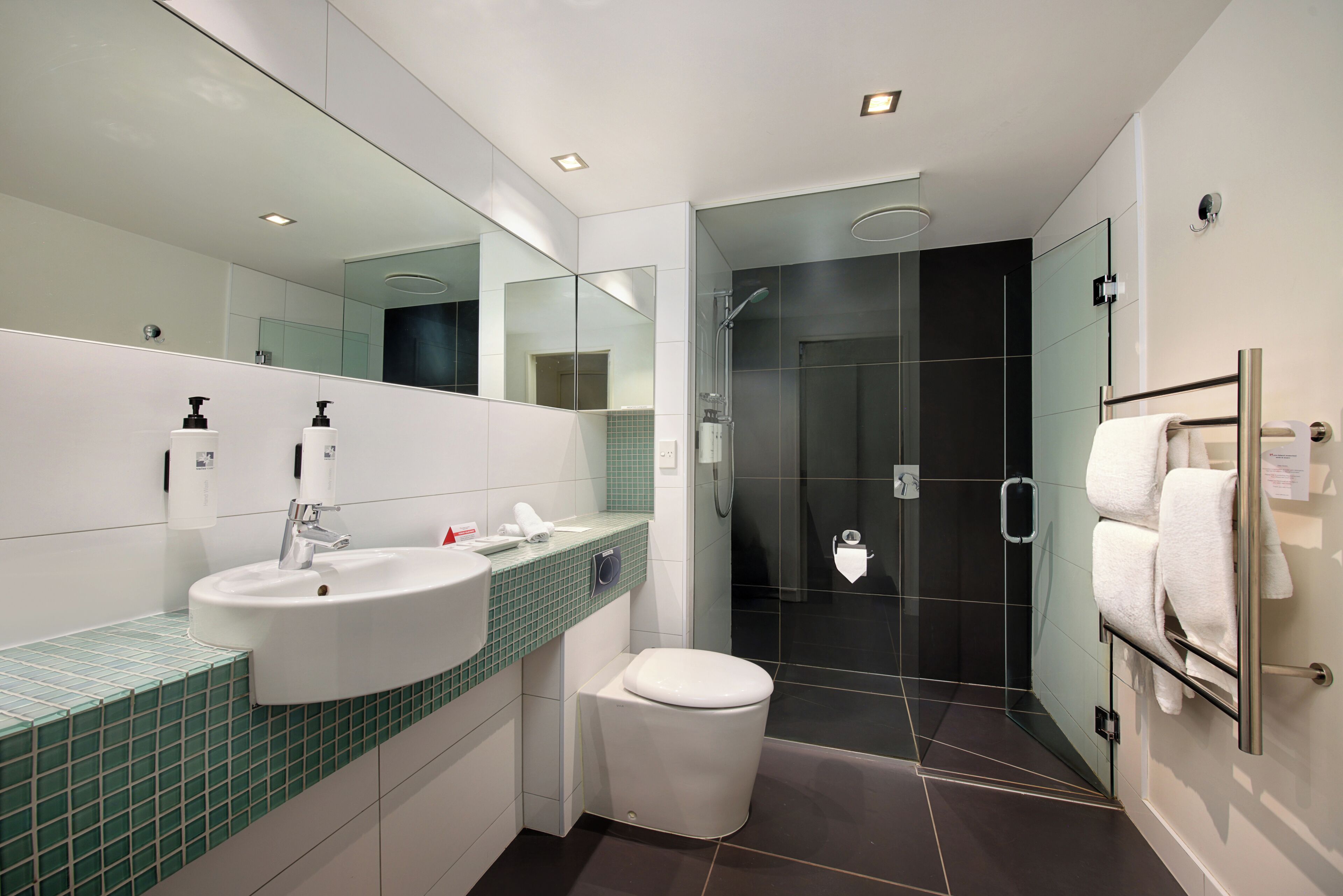 A modern bathroom with a white sink, green-tiled countertop, a toilet, a glass-enclosed shower, and a towel rack with towels.