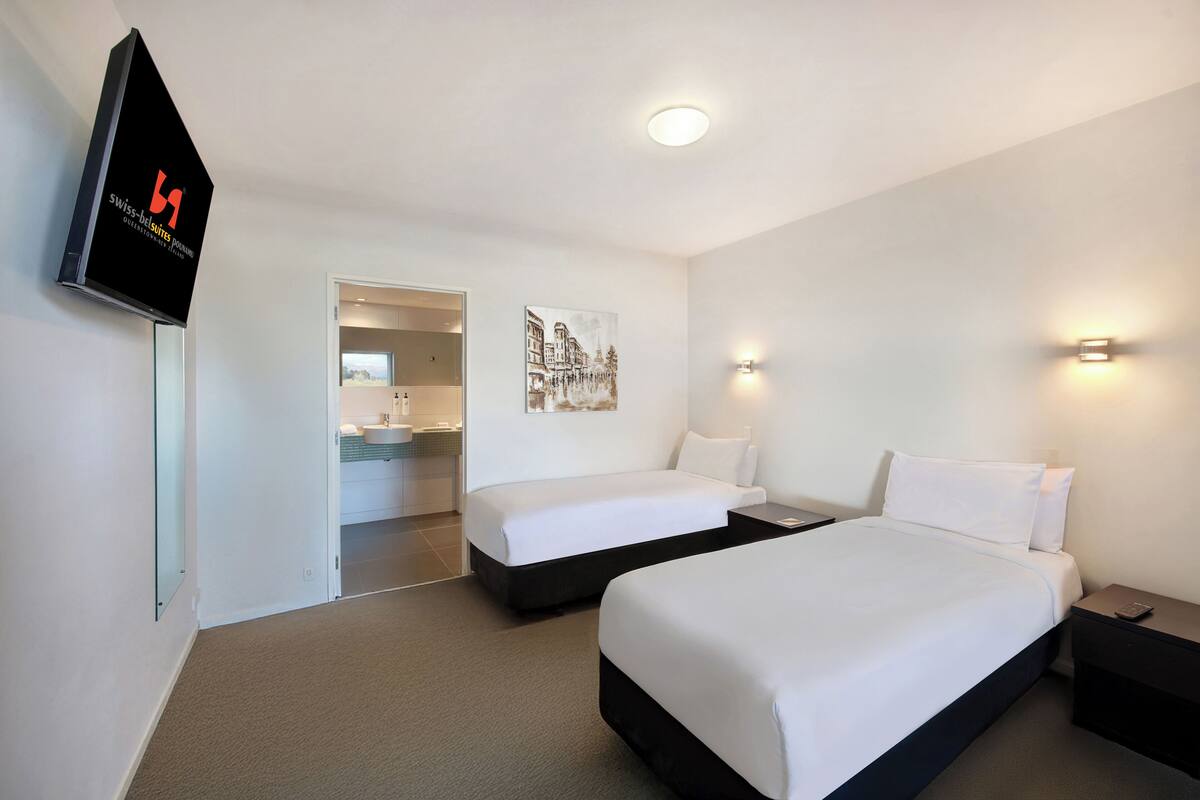 Habitación de hotel con dos camas, televisión, un cuadro y baño visible a través de una puerta abierta.