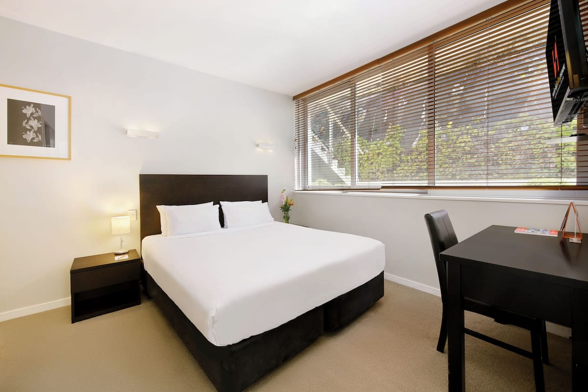 Habitación de hotel con cama, escritorio, silla, mesita de noche y ventana con persianas.