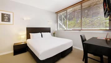 Habitación de hotel con cama, escritorio, silla, mesita de noche y ventana con persianas.