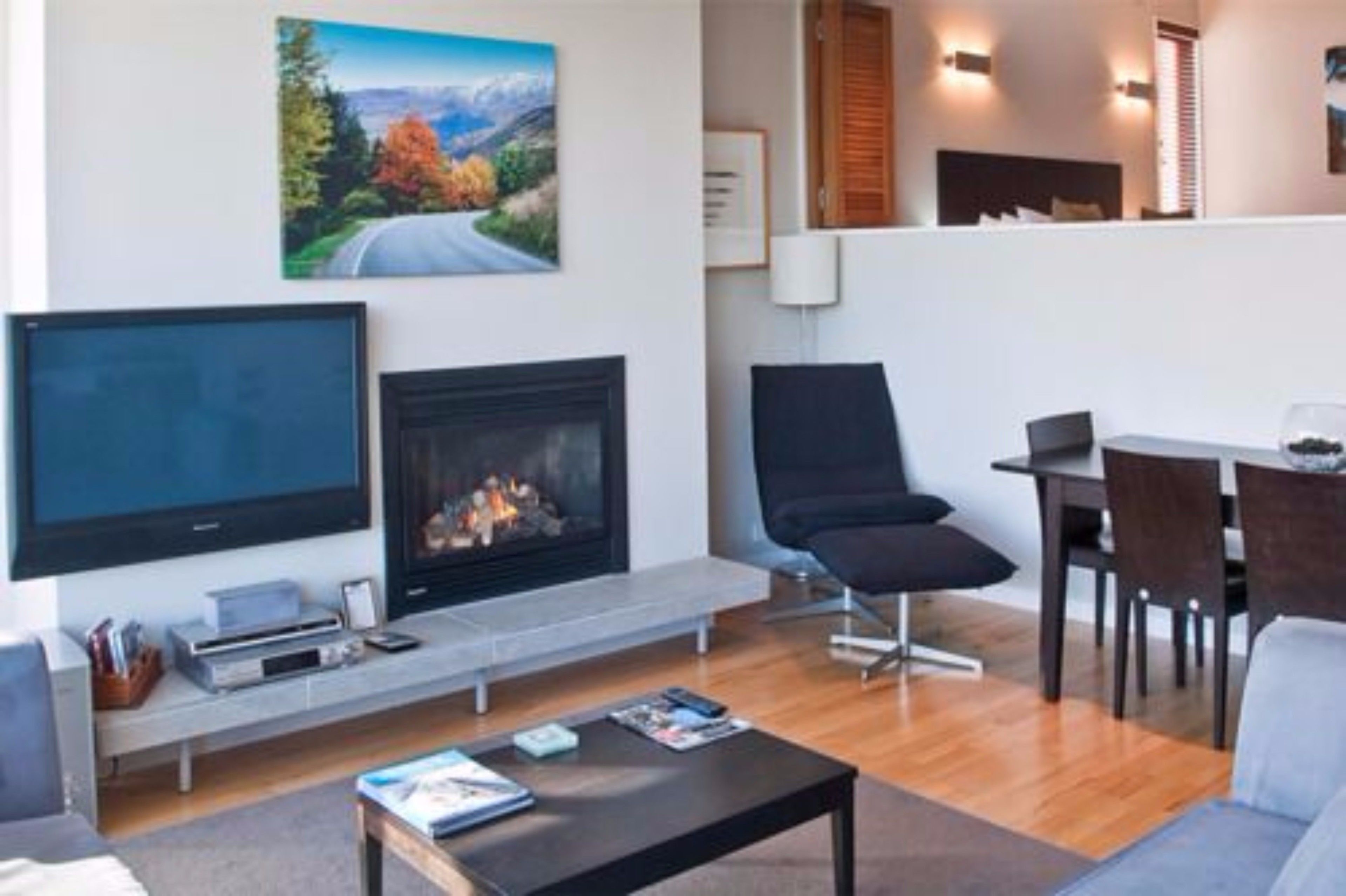 A modern living room with a fireplace, a flat-screen TV, a dining area with a table and chairs, and a painting of a scenic landscape on the wall.