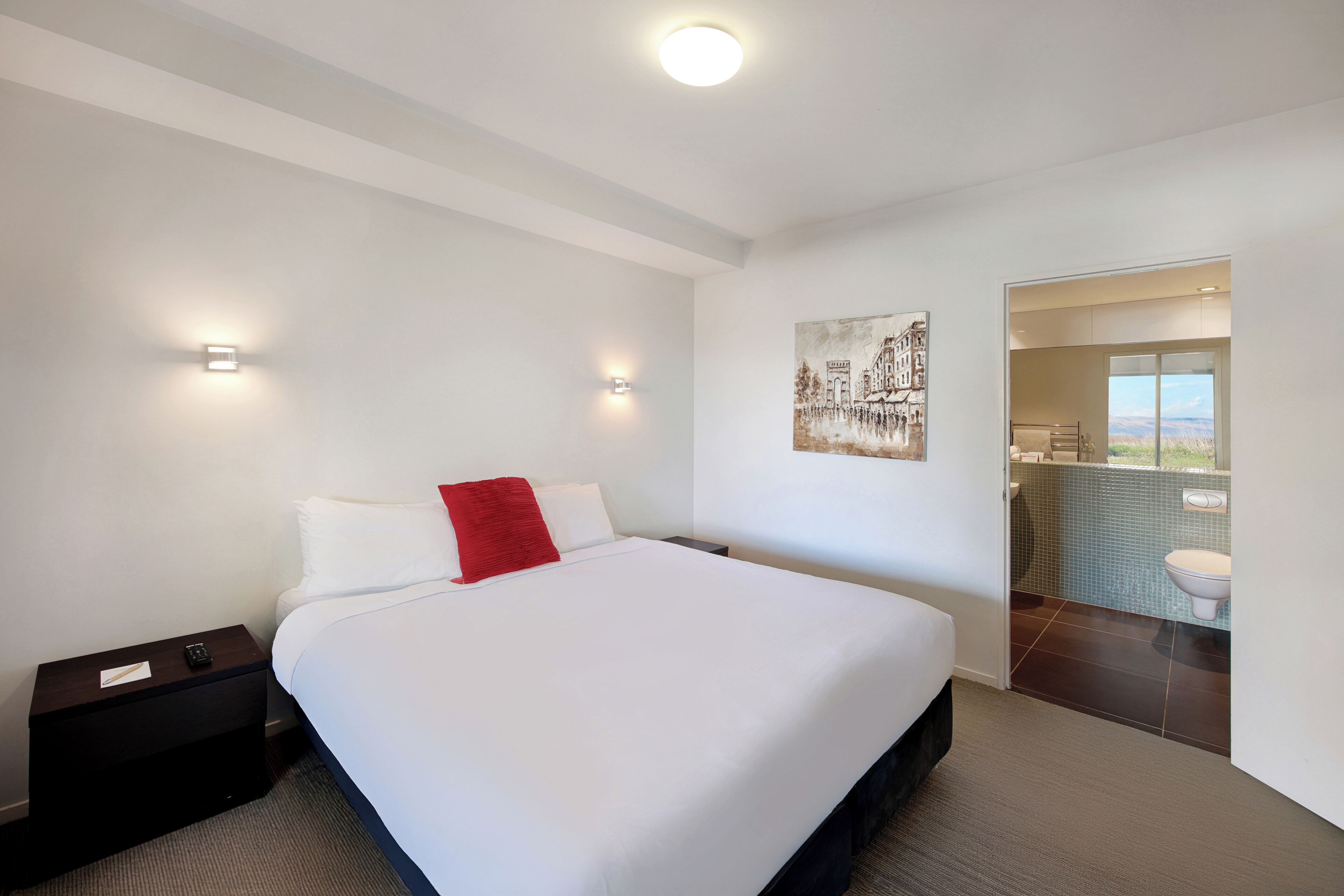 Habitación de hotel moderna con una cama grande, una mesita de noche, un cuadro en la pared y vistas al baño.