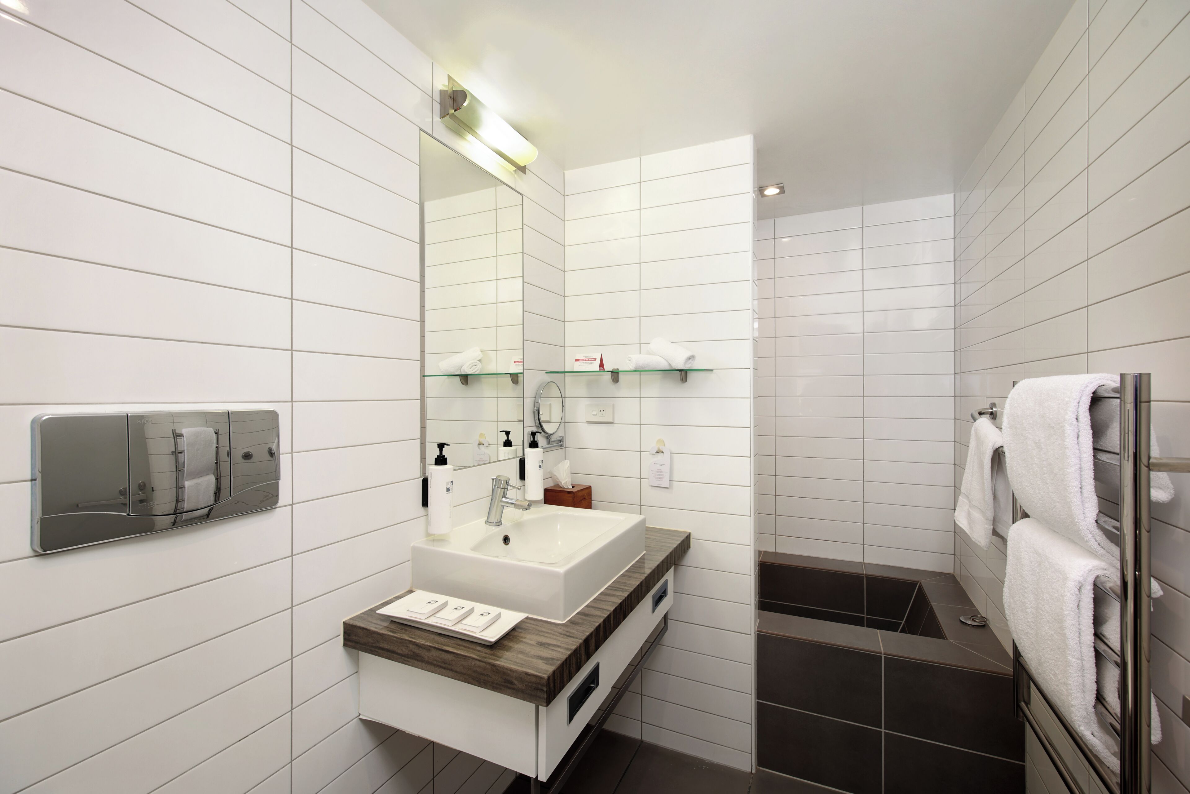 A modern bathroom with a white tiled wall, a wooden vanity, a large mirror, a built-in towel rack, and a separate shower area.