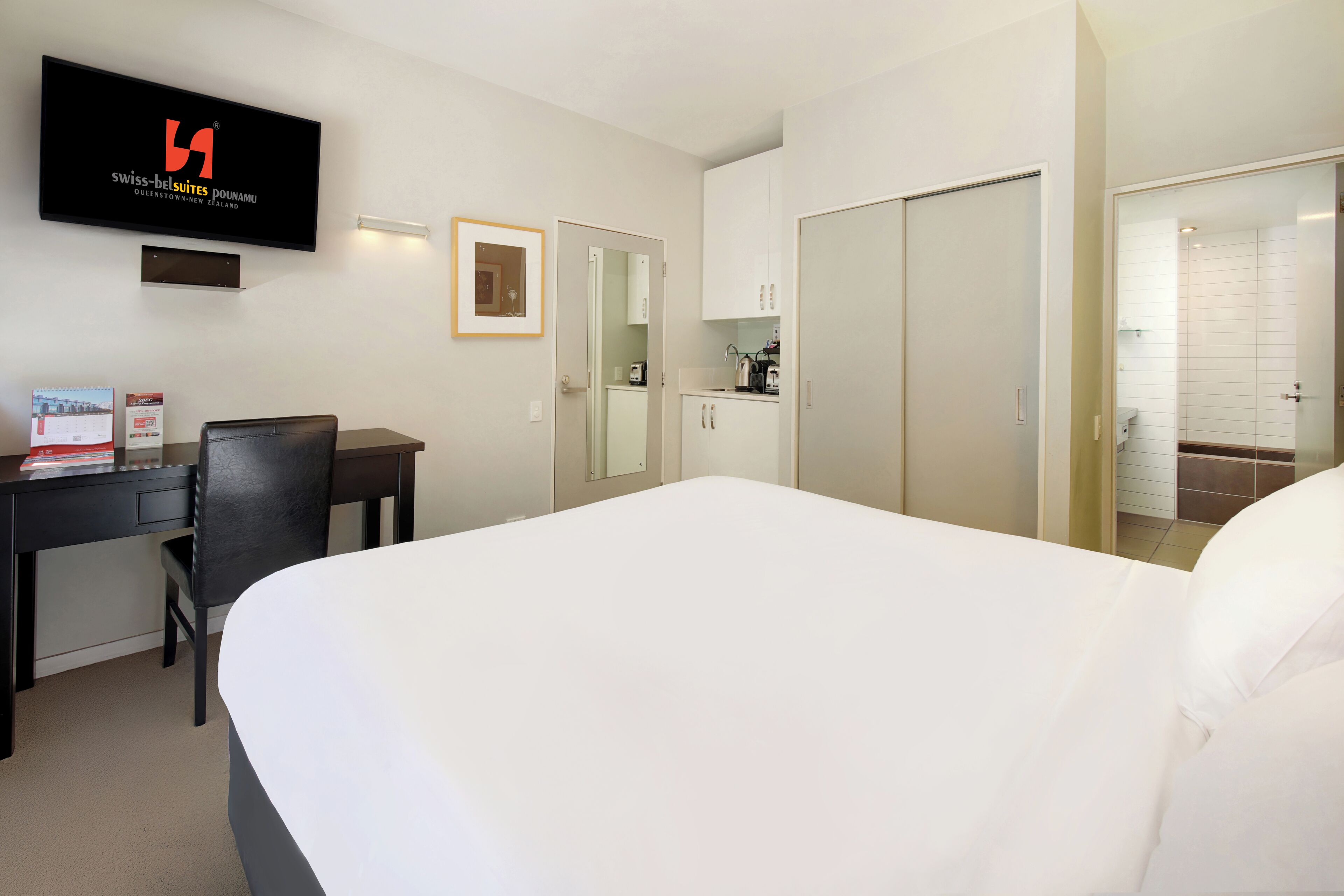 Et hotelværelse med seng, skrivebord, stol, fjernsyn, garderobeskab og et tekøkken.