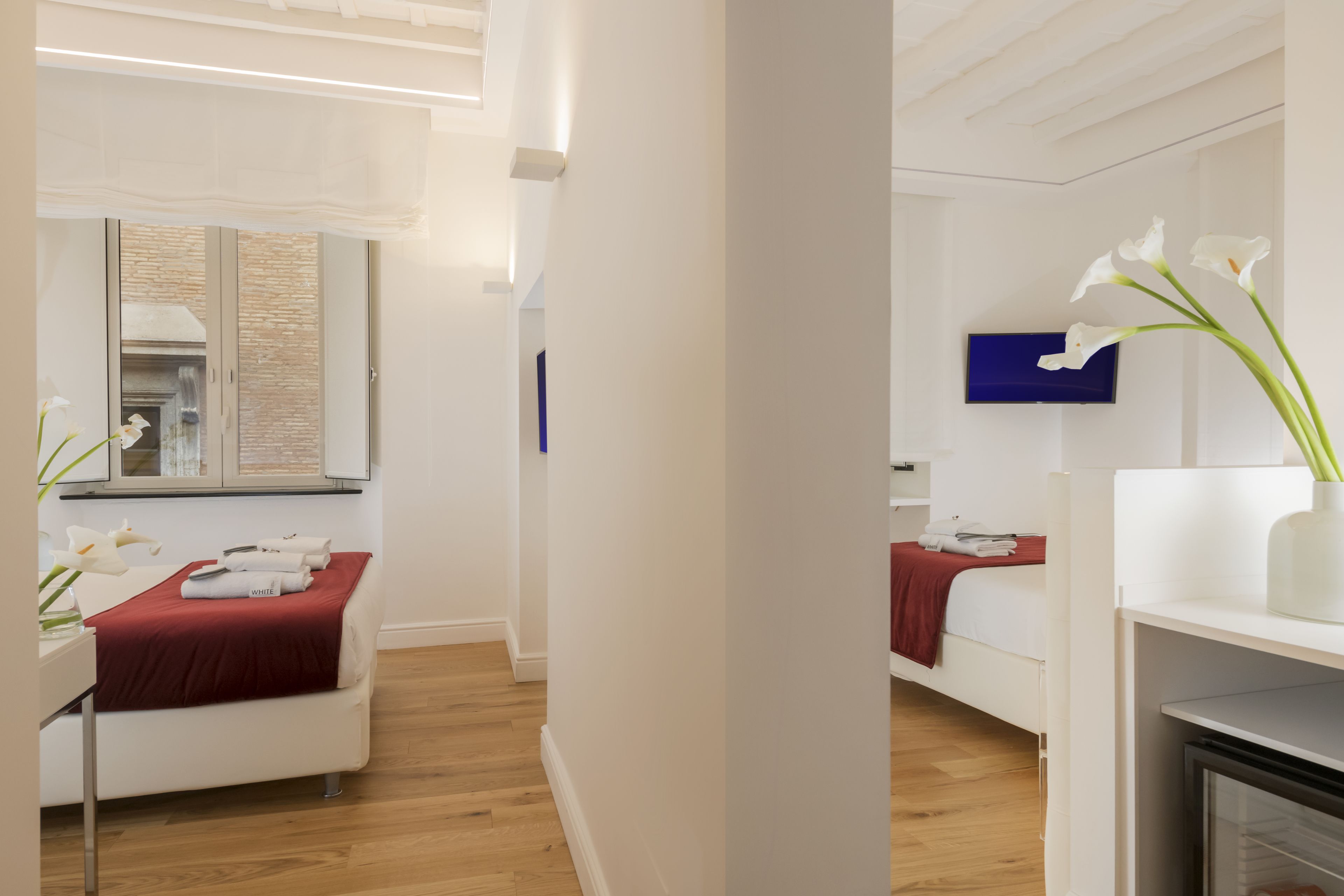 Une chambre d’hôtel moderne avec un plancher en bois, un lit recouvert d’une couverture rouge, une table de chevet blanche ornée d’un vase de fleurs blanches et un téléviseur à écran plat fixé au mur.