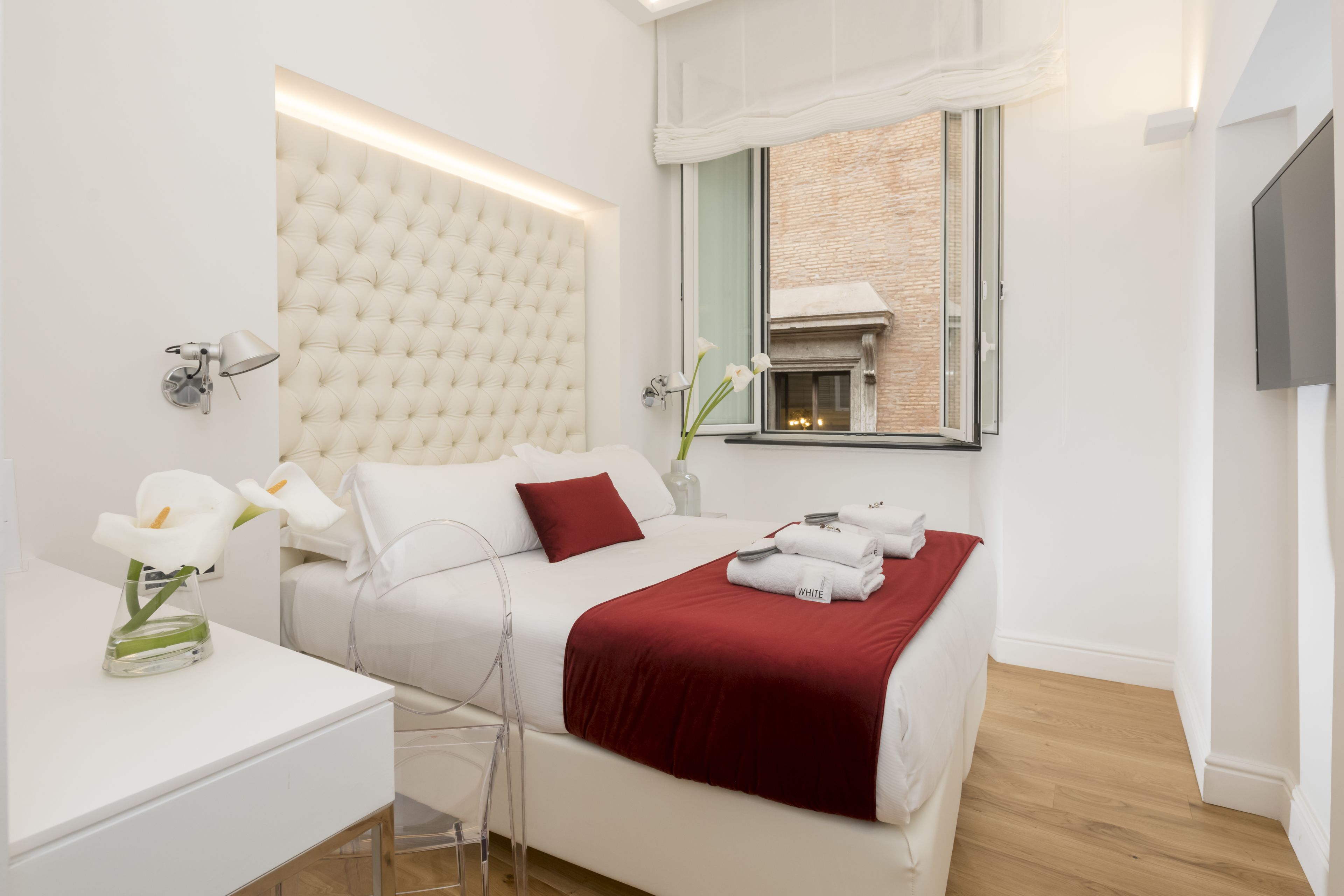 Quarto de hotel moderno com uma cama grande, roupa de cama branca, um cobertor vermelho, uma mesa de centro de vidro, um vaso com flores e uma lareira visível por uma janela.