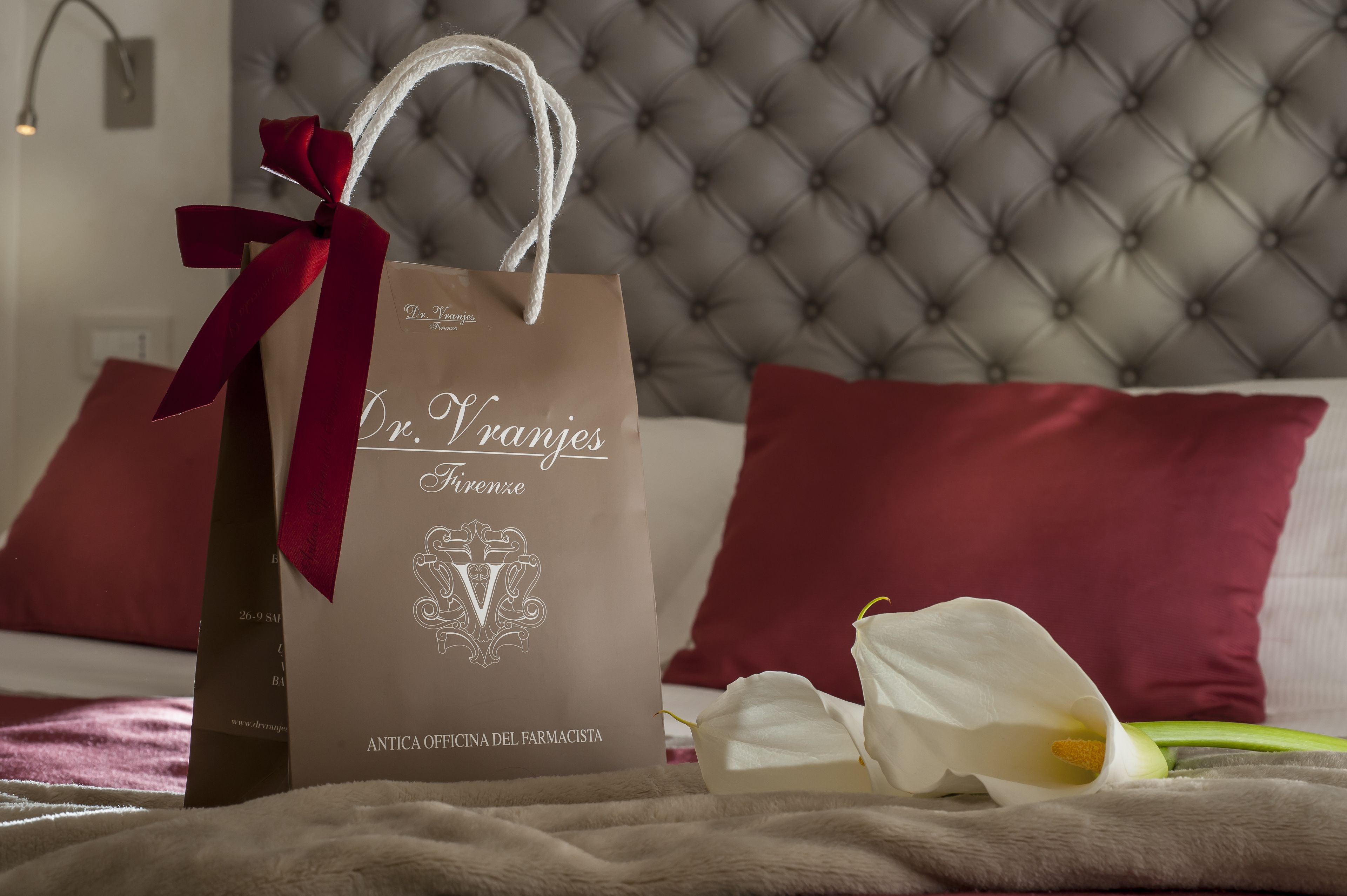 A gift bag with a red ribbon on a bed with a tufted headboard and red pillows.