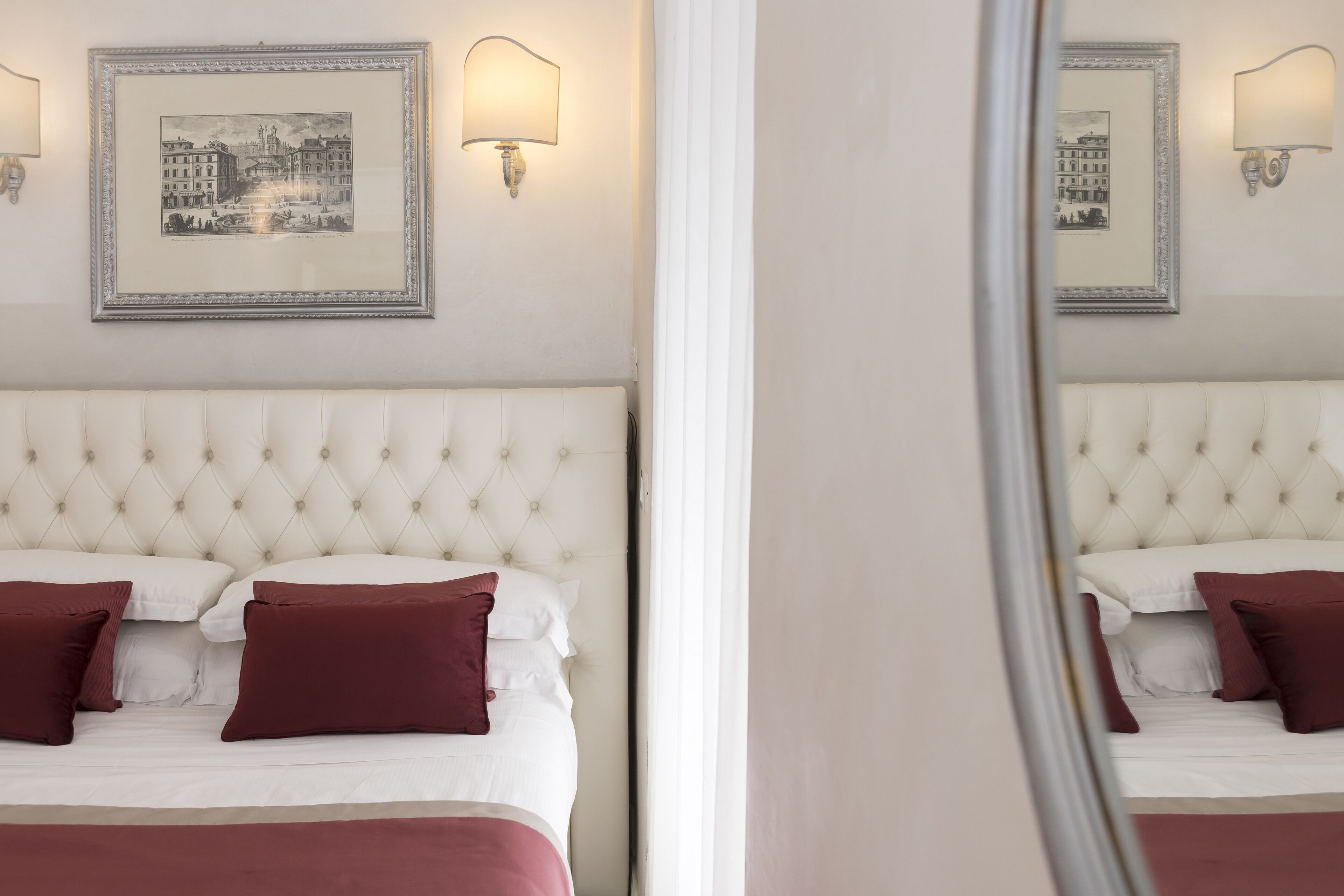 Una habitación de hotel con cabecera capitonada, cama con ropa de cama blanca y granate, dos lámparas fijadas a la pared y cuadros en la pared.