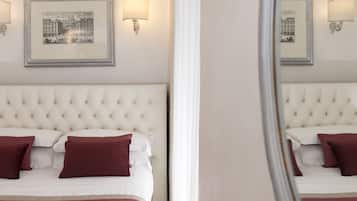 Una habitación de hotel con cabecera capitonada, cama con ropa de cama blanca y granate, dos lámparas fijadas a la pared y cuadros en la pared.