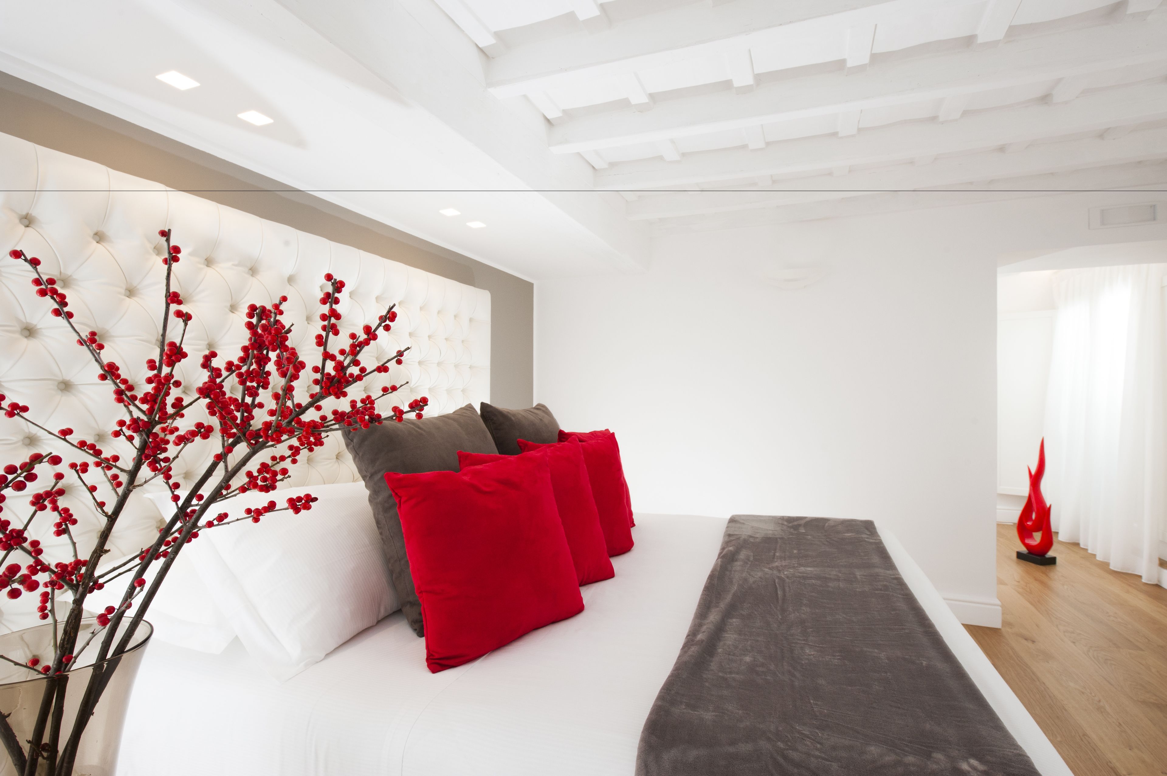 A modern hotel room with a tufted headboard, a bed with red and grey bedding, and a decorative red vase.