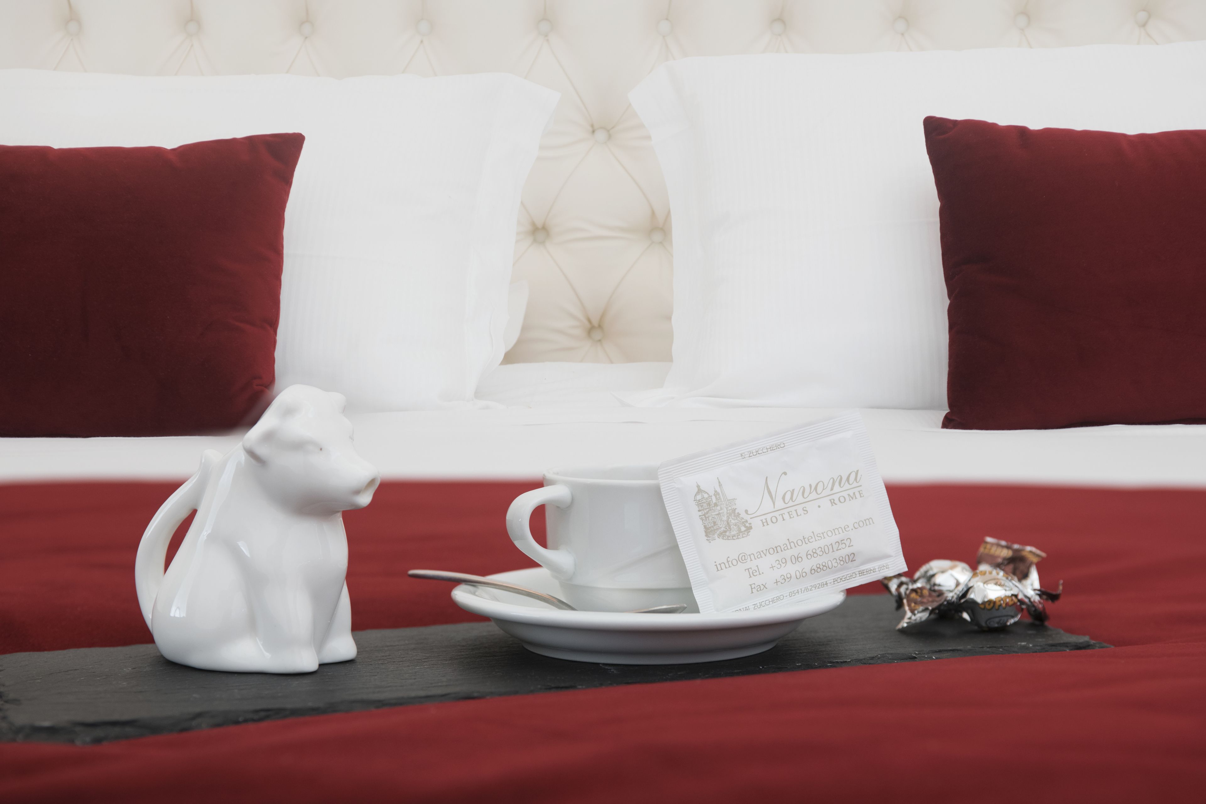 Un sofá blanco con cojines rojos, una figurita de perro de cerámica blanca, una taza y platillo blancos, y una tarjeta de presentación sobre un posavasos negro.