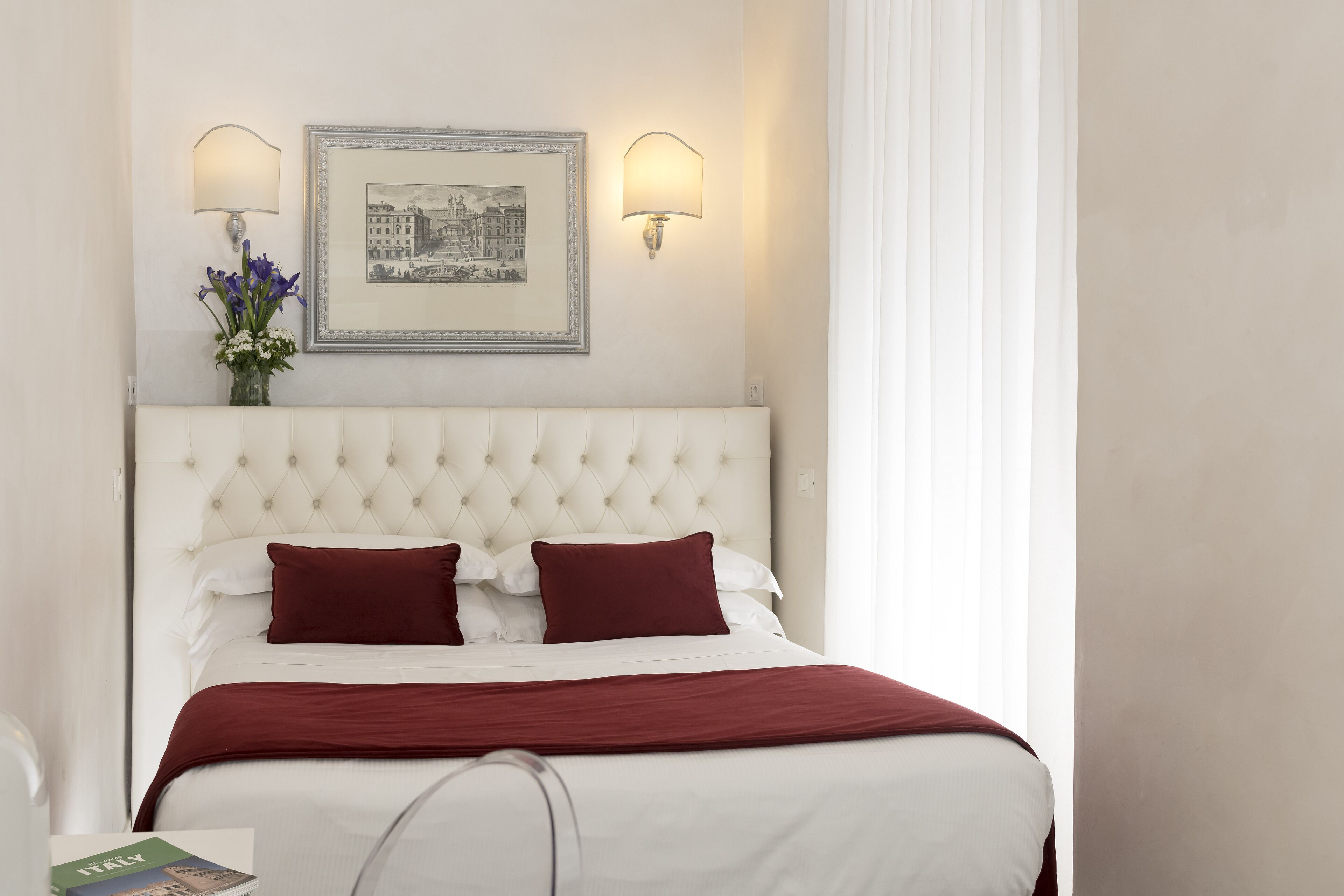 A hotel room with a tufted headboard, two wall-mounted lamps, a framed picture, a vase with flowers, and a red throw blanket.