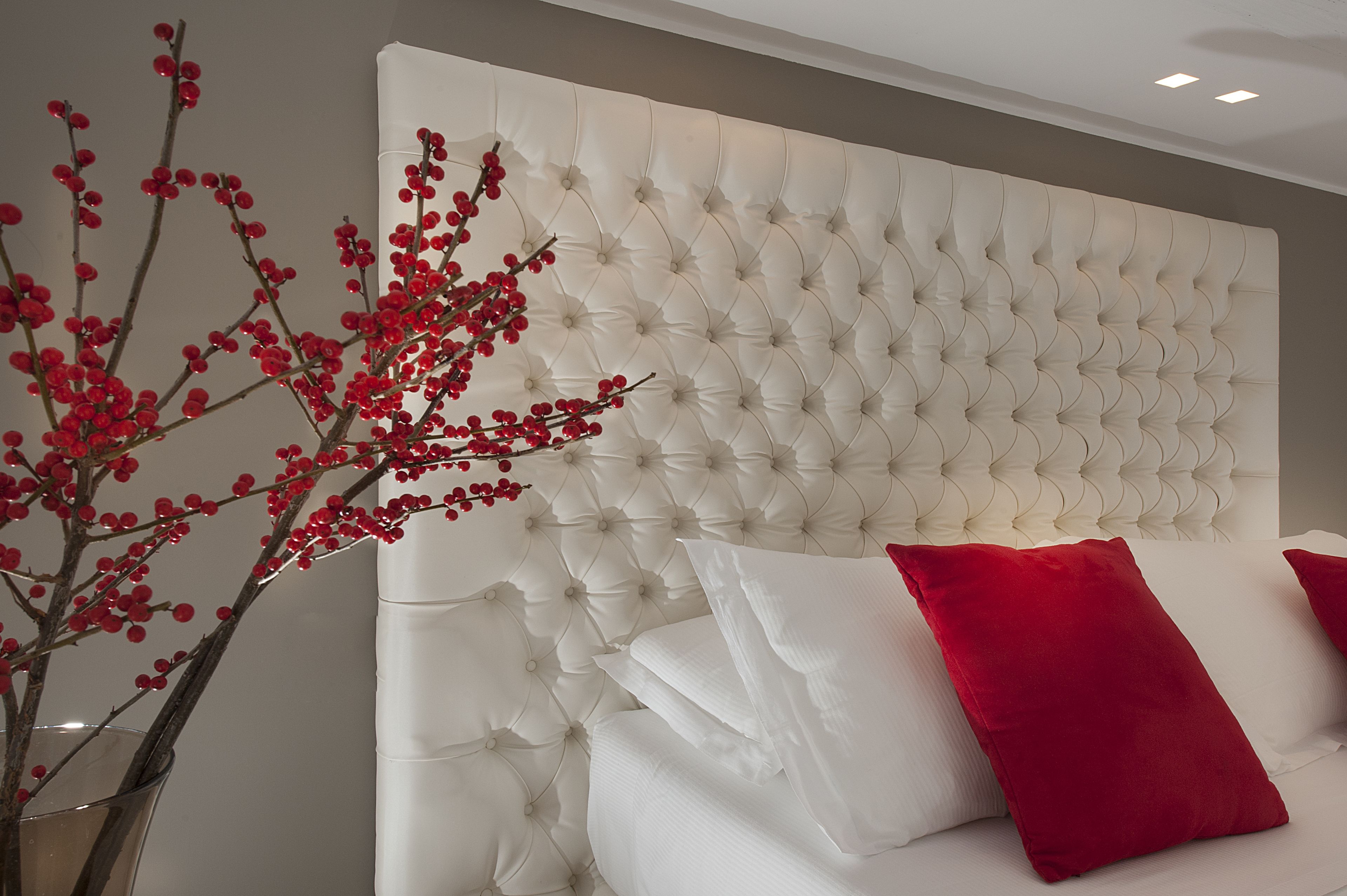 A bedroom with a tufted headboard, white bedding, and a vase with red berries.