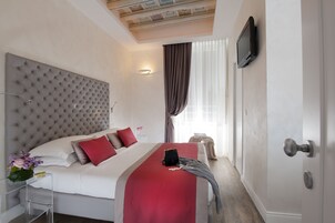 A hotel room with a tufted headboard, a bed with red and white bedding, a bedside table with a vase of flowers, and a television mounted on the wall.