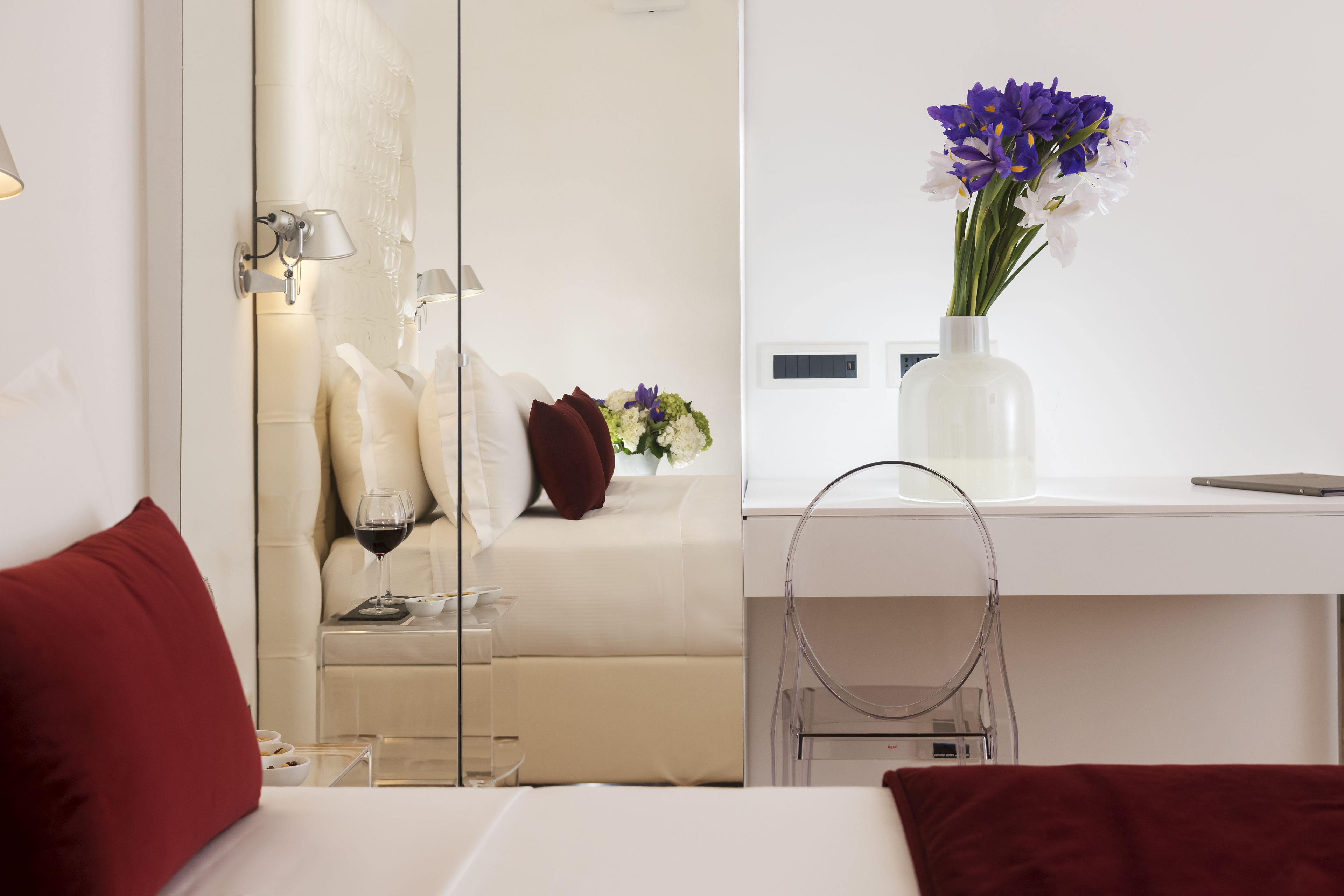 Habitación de hotel con cama, mesita de noche con una copa de vino, un jarrón con flores y un espejo que refleja la habitación.