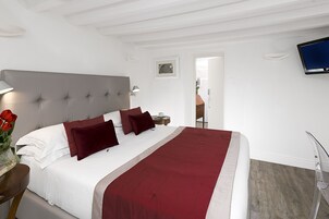 A hotel room with a large bed, a bedside table with a lamp, a TV mounted on the wall, and a view of the outside through an open door.