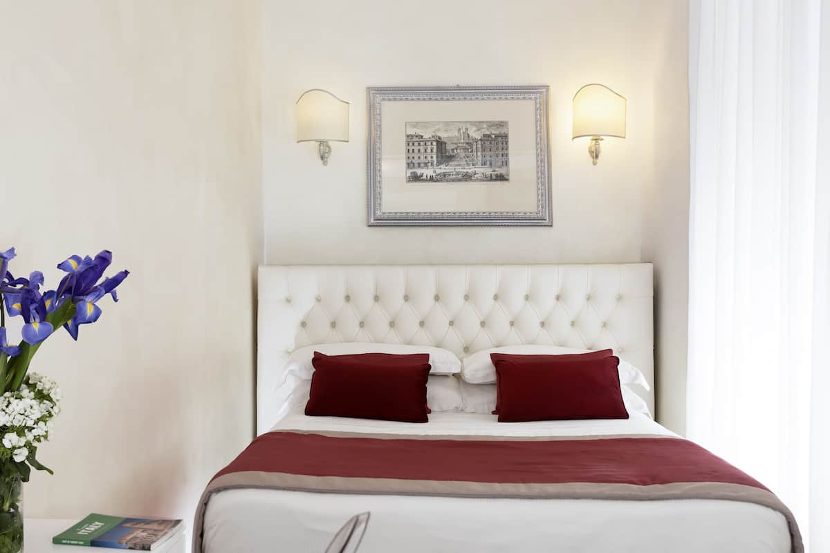 A neatly made bed with a white headboard and red pillows, a framed picture on the wall, and a vase of flowers on a nightstand.