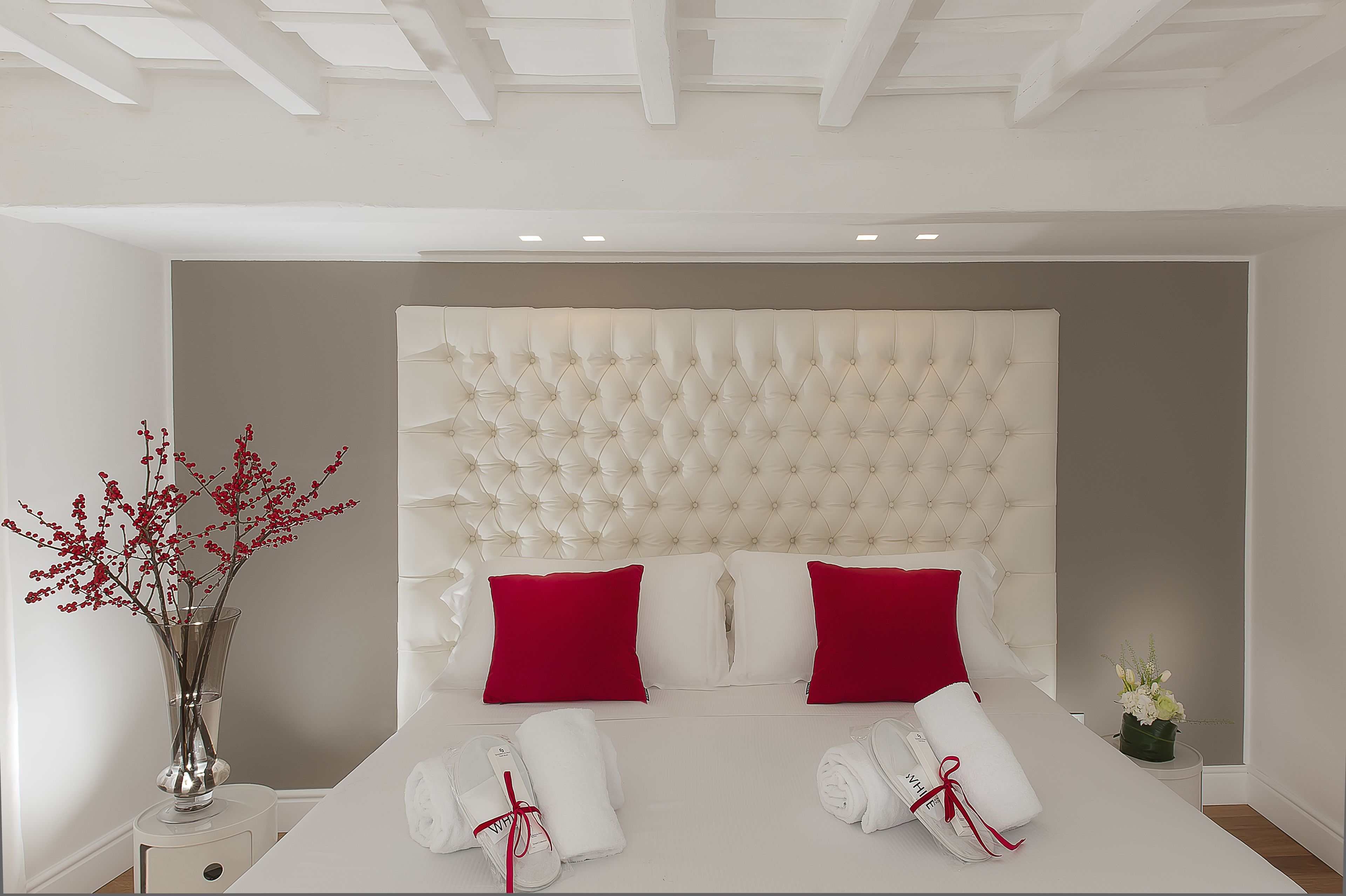 A neatly arranged hotel room with a tufted headboard, red and white pillows, a vase with red flowers, and neatly folded towels.