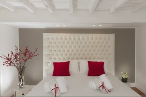 A neatly arranged hotel room with a tufted headboard, red and white pillows, a vase with red flowers, and neatly folded towels.