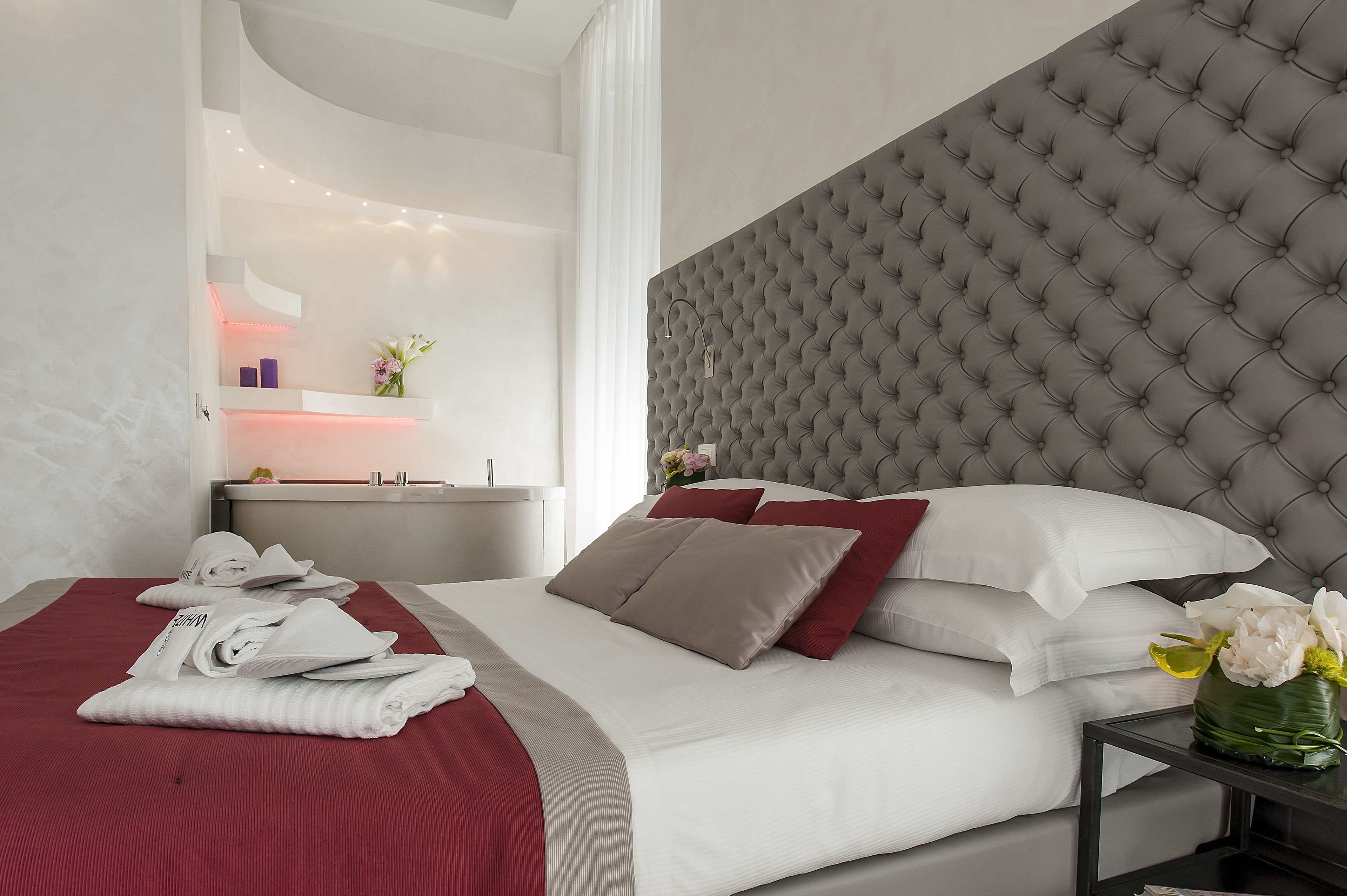 Una habitación de hotel moderna con una cama grande, un cabecero capitonado gris y una mesita de noche con un jarrón de flores.