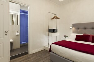 A bedroom with a bed, a bedside table, a nightstand, a mirror, a hanging coat rack, and a bathroom with a shower and toilet.