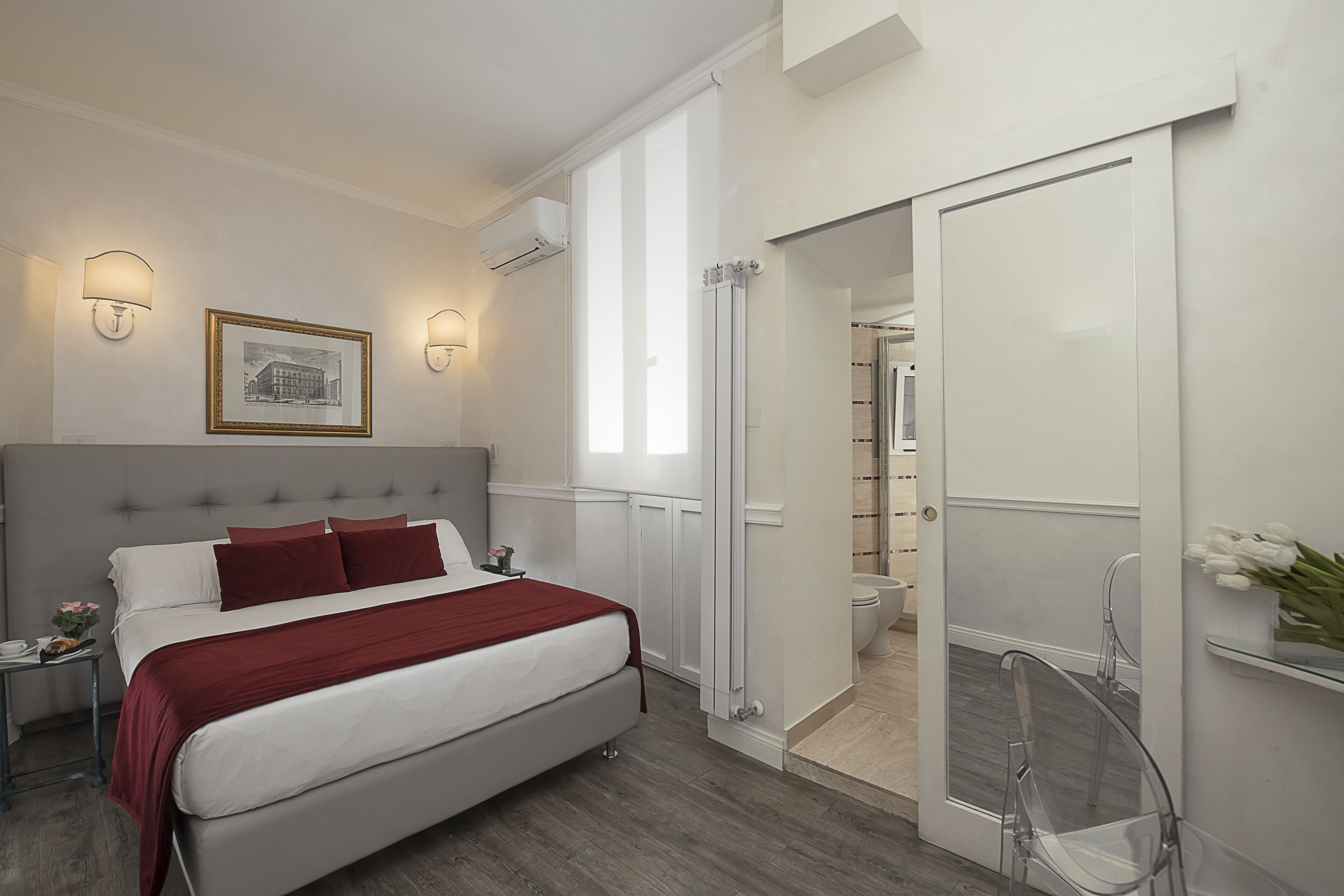 Un dormitorio moderno con una cama grande, una mesita de noche, un cuadro enmarcado y un baño visible a través de una puerta abierta.