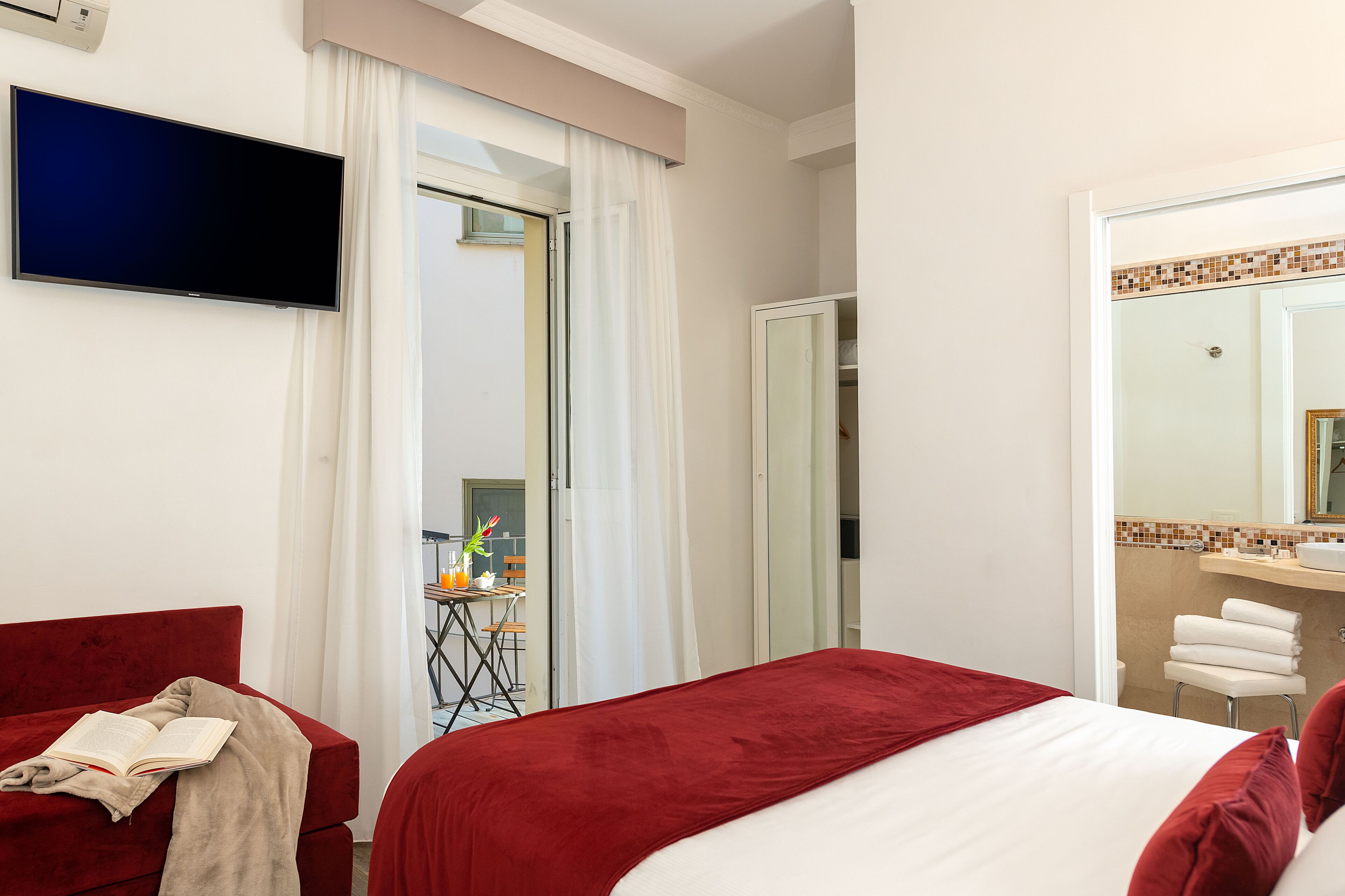 A hotel room with a bed, a red sofa, a TV, a bathroom with a mirror, and a small table with a plant.