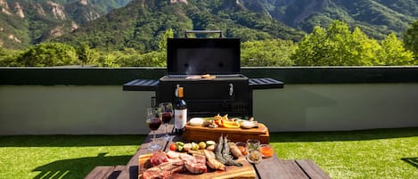 An outdoor barbecue setup with a grill, meats, vegetables, and drinks on a wooden table, overlooking a mountainous landscape.
