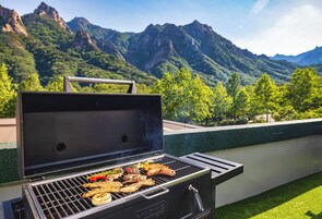 A barbecue grill with various meats and vegetables, set against a mountainous backdrop.