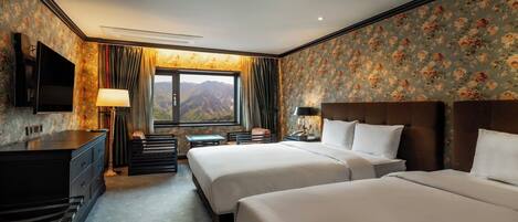 A hotel room with two beds, a desk, a TV, and a window with a mountain view.