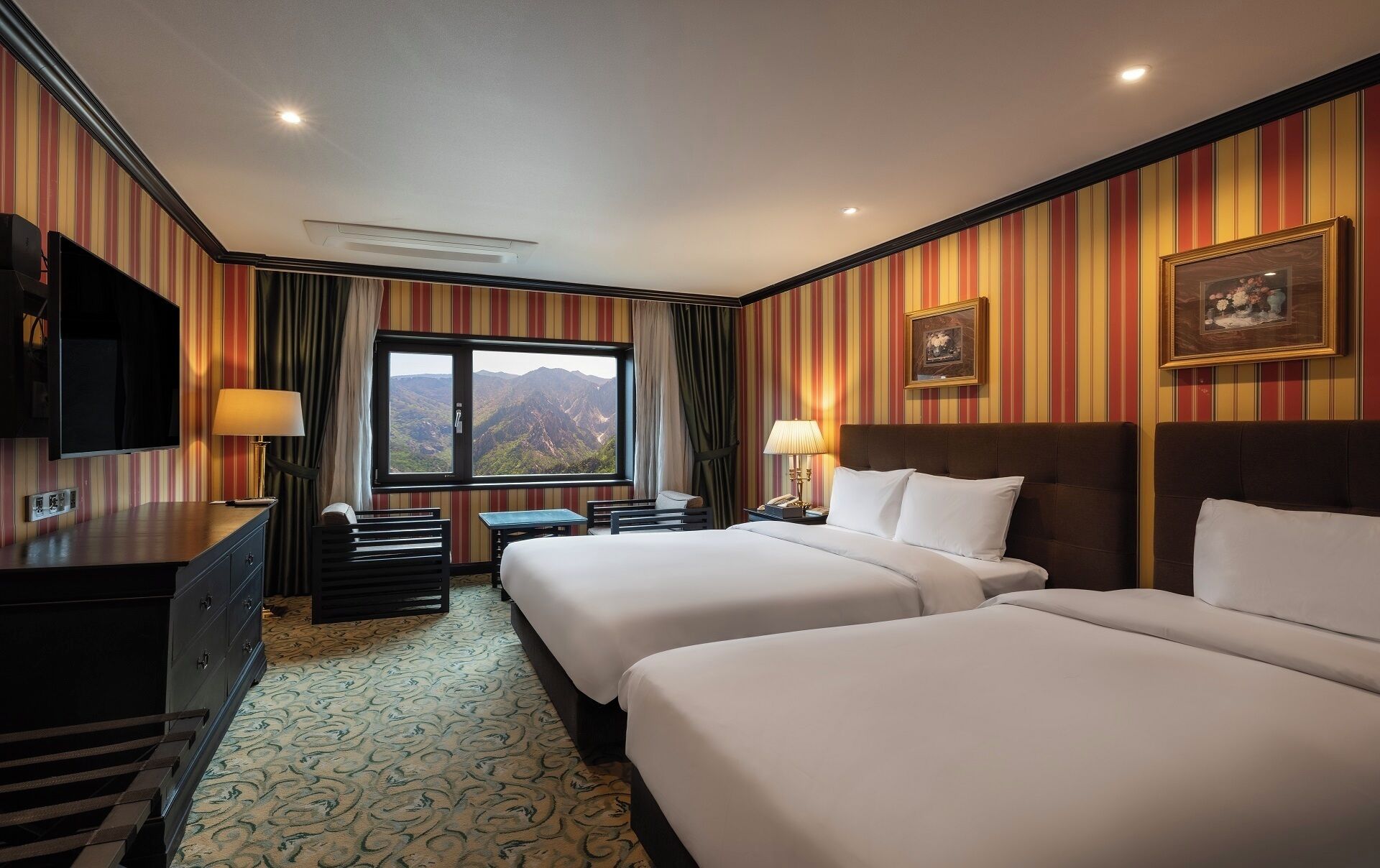 A hotel room with two beds, a desk, a TV, a window with a mountain view, and striped wallpaper.