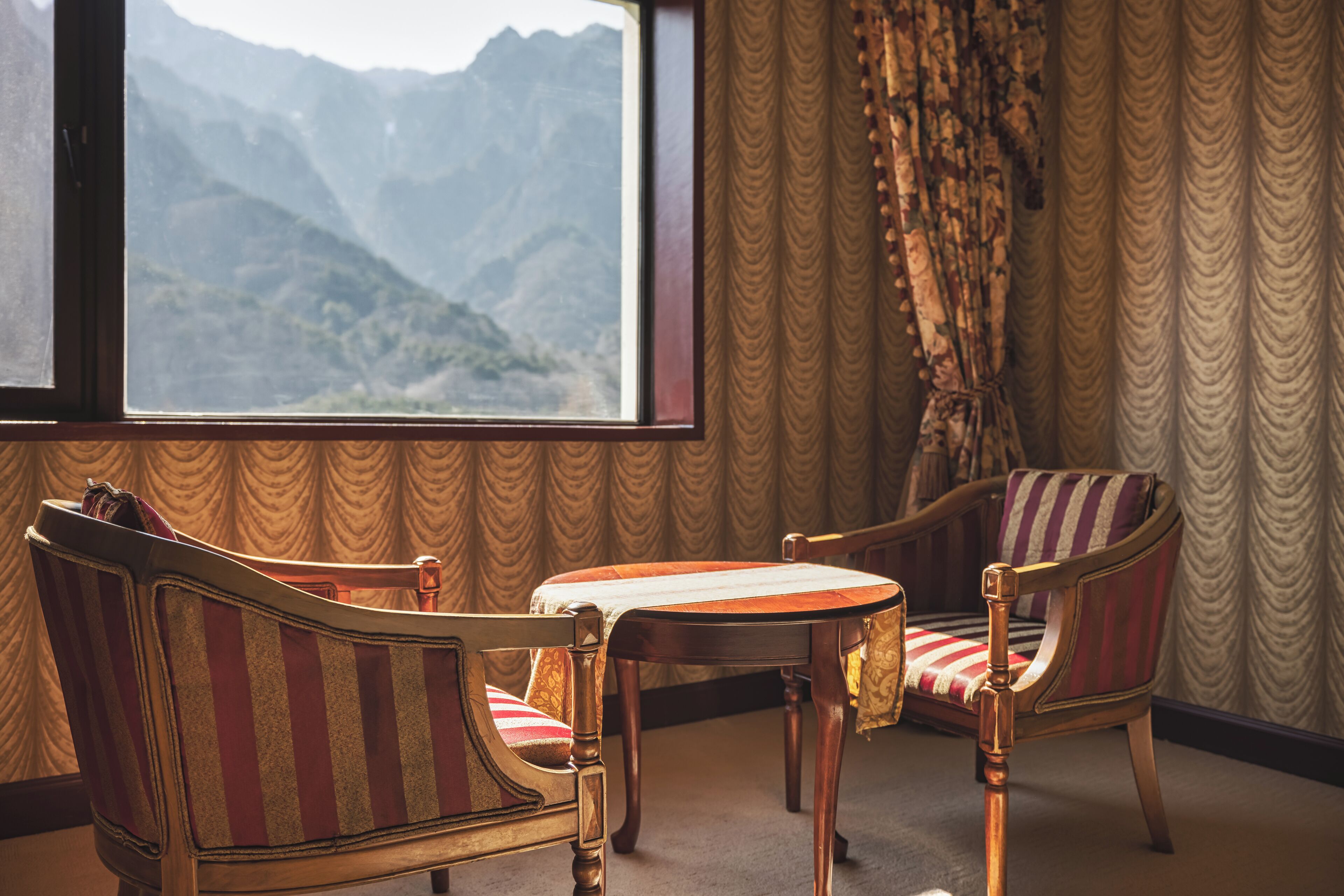 Habitación con una ventana grande que ofrece vistas a la montaña, dos sillones tapizados con estampado a rayas y una pequeña mesa de madera.