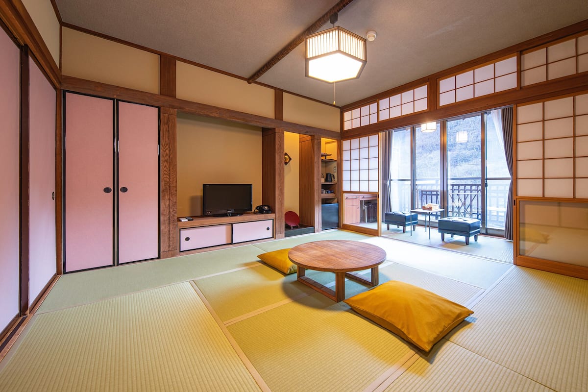 Een kamer met tatami matten, een tatami vloer, een lage houten tafel en kussens. Er staat een televisie op een lage houten kast, en een schuifdeur die leidt naar een andere kamer.