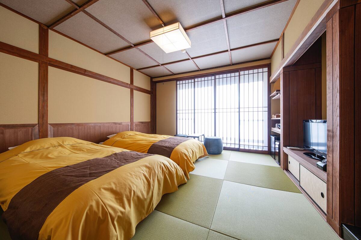 Een traditionele Japanse kamer met tatami vloeren, een bed met geel en bruin beddengoed, een televisie op een houten kast en een raam met verticale jaloezieën.